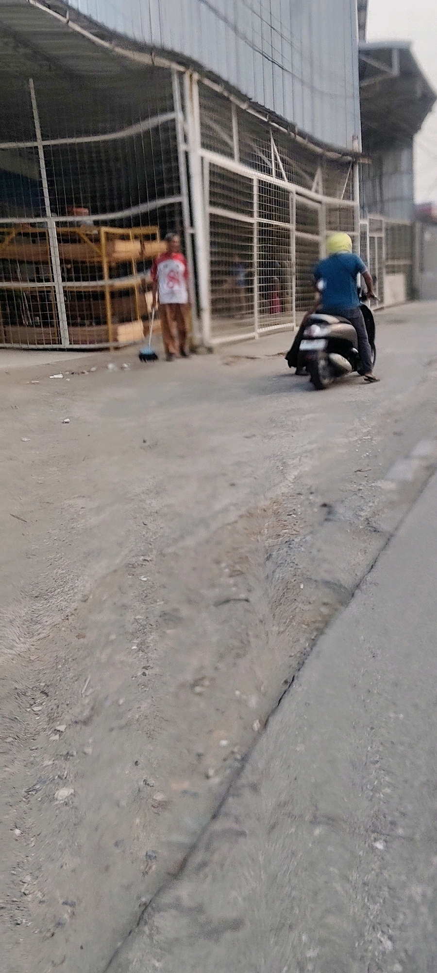 In Mekarmukti, Indonesia, a person wearing a blue shirt and a yellow helmet is riding a scooter on a paved road. The scooter is dark colored and appears to be a scooter. The person is facing away from the camera. To the left, another person wearing a red and white shirt and brown pants is sweeping the ground with a broom. This person is also facing away from the camera. Behind the people, there is a building with a metal roof and white metal fencing. The building appears to be a warehouse or storage facility. Wooden shelves are visible inside the fenced area, with some items stored on them. The ground is a mix of asphalt and dirt. The overall scene depicts an outdoor area with commercial or industrial structures and activity.