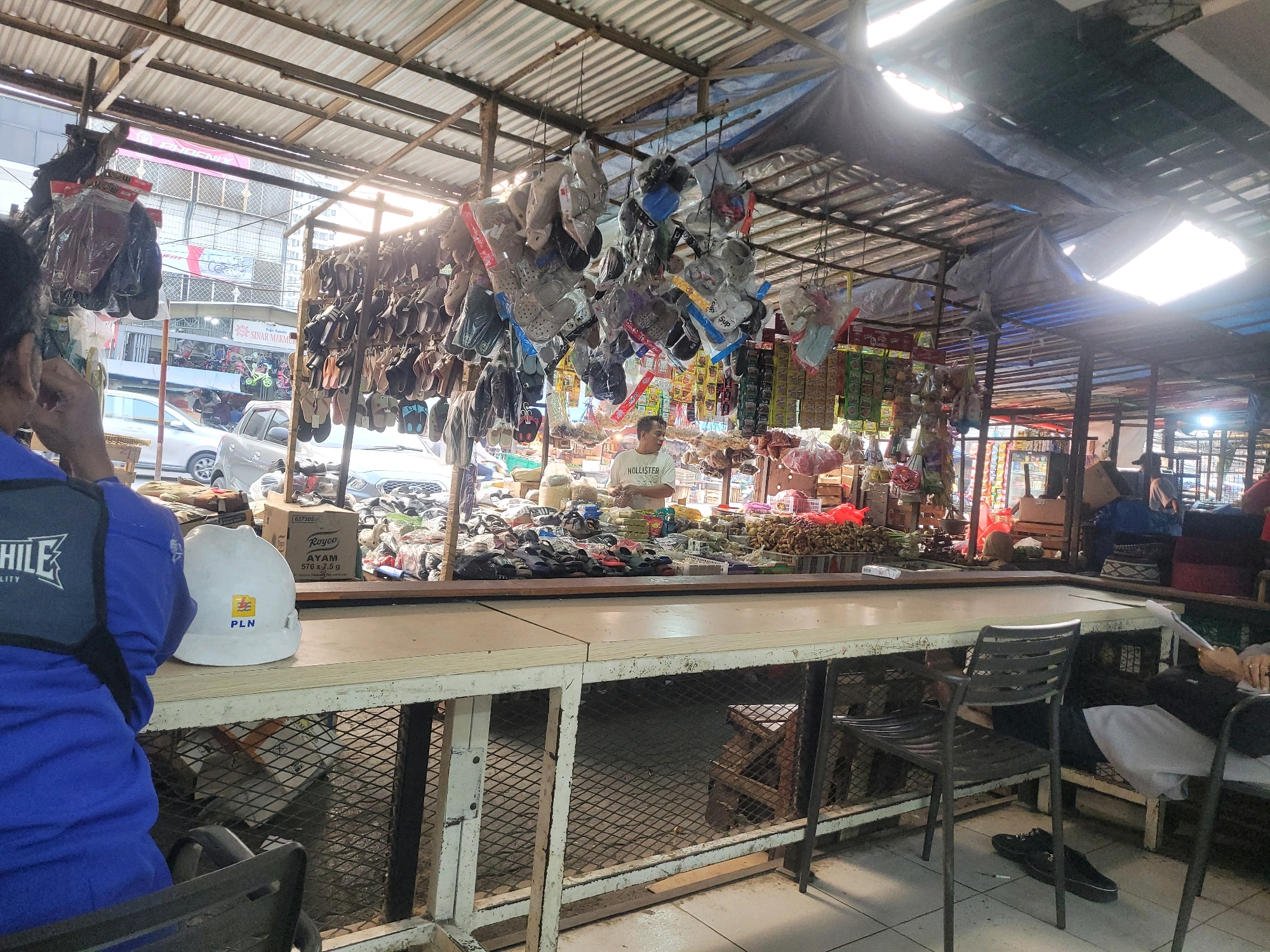 The image captures an outdoor market scene in South Jakarta, Indonesia. The foreground features a table with a white surface and a metal mesh base, where a white hard hat with a "PLN" logo is placed. To the left, a person wearing a blue uniform vest is partially visible. Behind the table, a stall is set up with various items for sale, including footwear hanging from the ceiling, bags of snacks or spices, and other goods. A man in a white t-shirt stands behind the stall, interacting with the environment. The market is covered by a corrugated metal roof with some tarpaulin, allowing sunlight to stream through gaps. In the background, vehicles and buildings suggest a bustling urban setting. The overall atmosphere is that of a typical Indonesian street market.