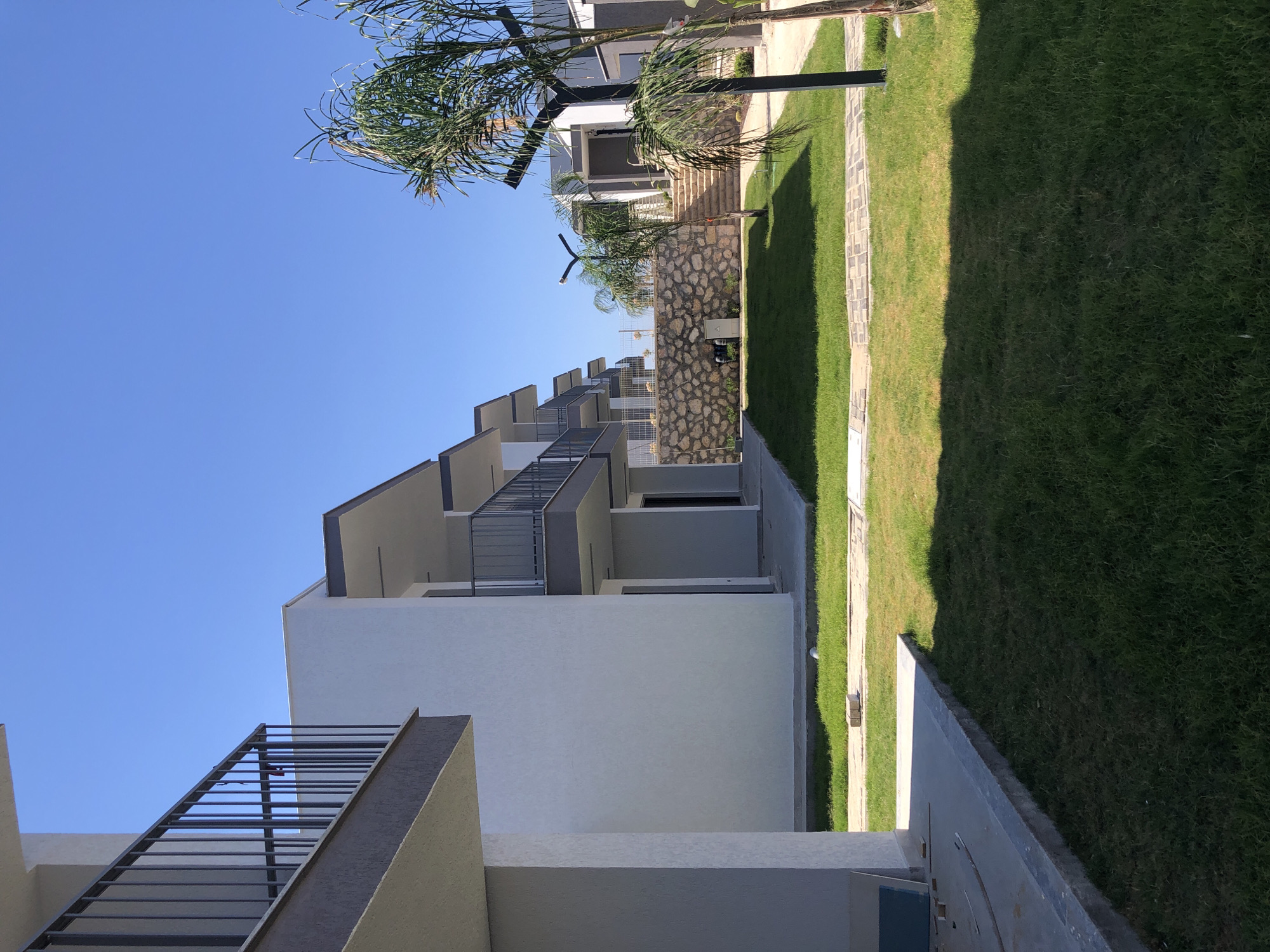 This is a photograph of an outdoor courtyard area in Karaburun, Turkey, likely a residential complex or hotel. The scene is dominated by modern, multi-story white buildings with dark trim and balconies. The architecture is clean and minimalist. A paved walkway runs along the front of the buildings, bordered by a well-maintained, green lawn. Beyond the lawn, there is a decorative stone wall. Steps lead up from the courtyard towards another section of the complex, where more buildings are visible. A palm tree stands in the foreground, casting a shadow on the grass. The sky is a clear, bright blue, suggesting a sunny day. There are no people or animals visible in the image. The overall impression is one of a newly developed or well-kept outdoor space.