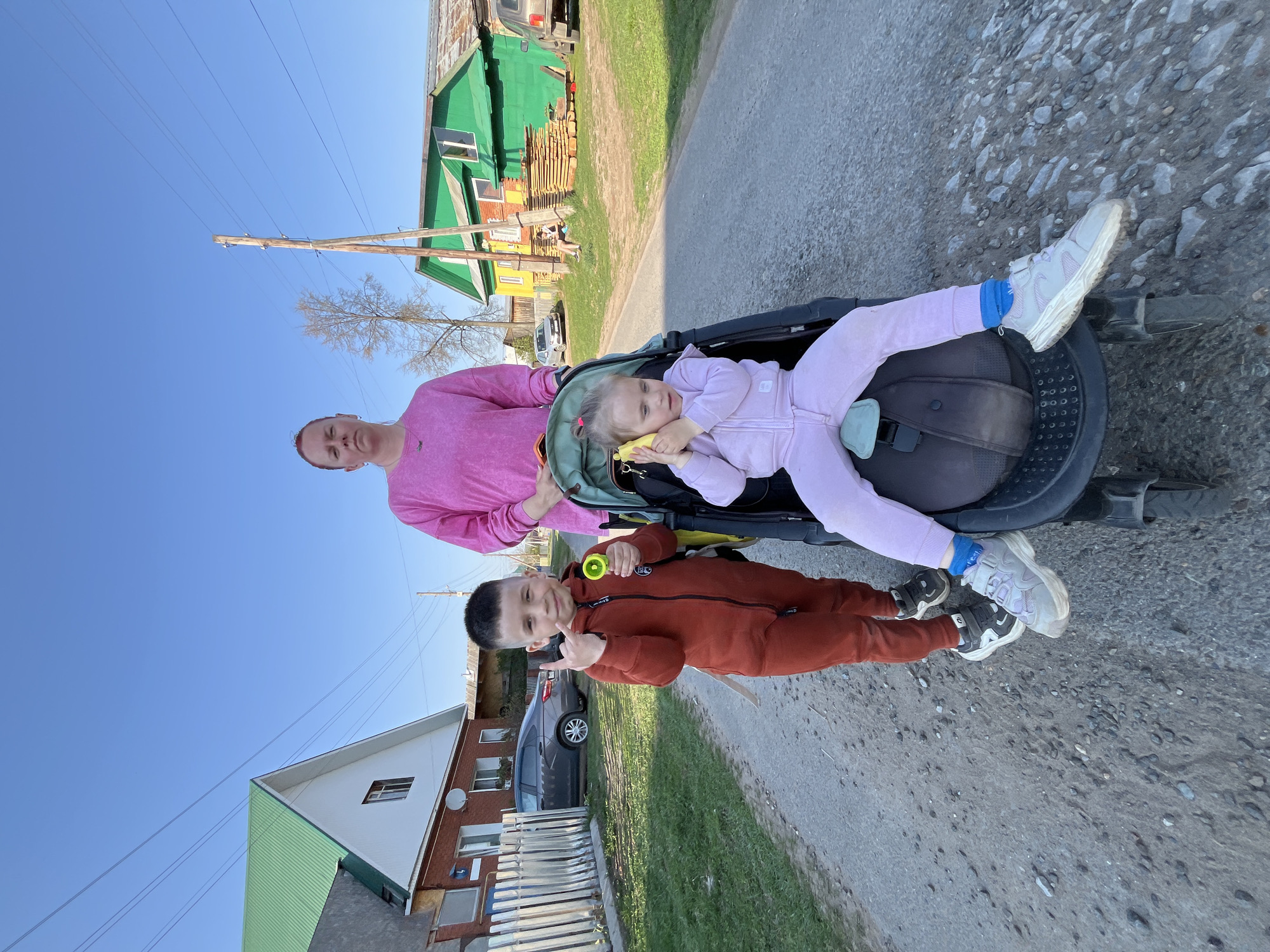 On a sunny day in Chaikovsky, Russia, a woman in a pink shirt stands on a paved road next to a black stroller. Inside the stroller lies a child dressed in a lavender jumpsuit. A boy in an orange jumpsuit stands in front of the stroller holding a yellow toy. Behind the woman is a green and yellow house. Across the road, there is a white and brown house with a dark grey car parked in front. The sky is clear and blue.
