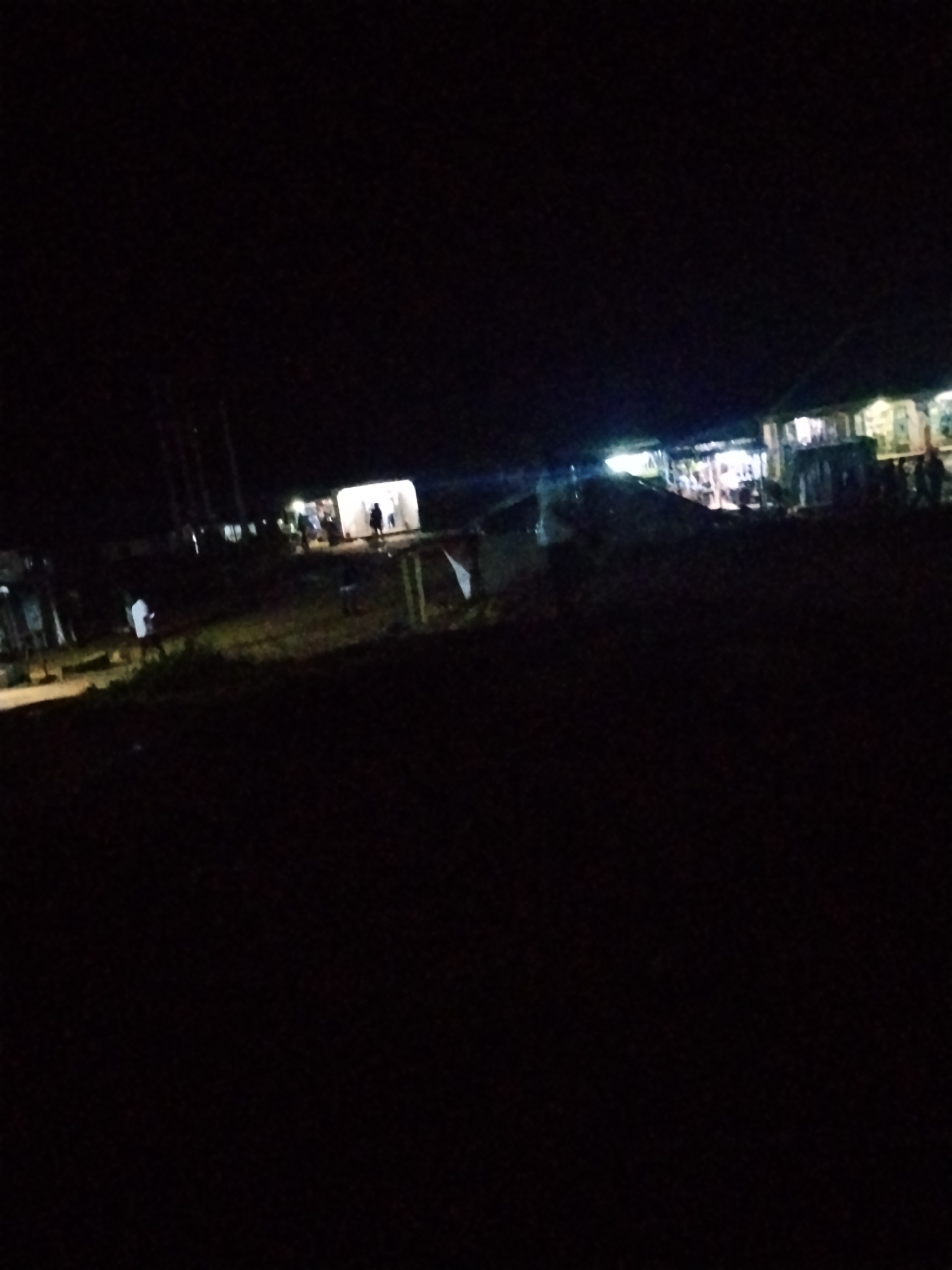 The image depicts a dimly lit outdoor scene at night in Durumi, Nigeria. Several buildings are visible, illuminated by artificial lights, suggesting they are businesses or residences. People are present in the scene, some walking and others gathered near the buildings. The overall impression is of a community gathering or activity taking place after dark.
