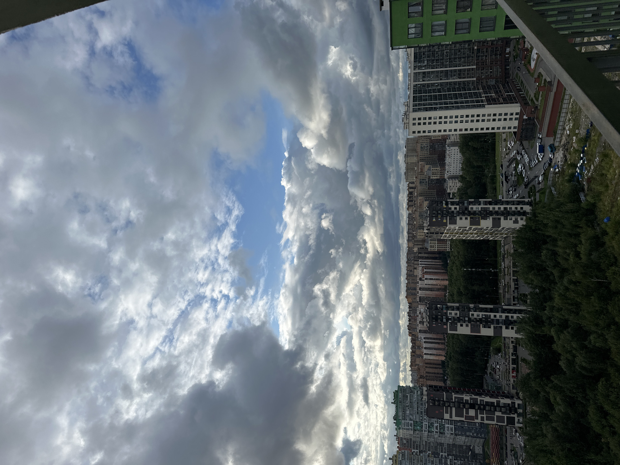 The image depicts a dense urban landscape with numerous high-rise buildings and a thick canopy of trees under a dramatic sky filled with heavy, gray clouds interspersed with patches of blue. In the foreground, a metal railing is visible, suggesting the viewpoint is from a balcony or elevated structure.
The cityscape features a variety of apartment complexes, characterized by their diverse architectural styles and color palettes, ranging from muted browns and beiges to brighter greens and whites. Two prominent, tall, dark-patterned buildings stand out in the mid-ground. A large expanse of dark green trees separates the foreground from the distant buildings. The sky is predominantly overcast, with sunlight breaking through in some areas, creating a sense of depth and dynamic lighting. There are no visible people in the scene, and the focus is on the urban environment and the atmospheric conditions. The scene conveys a sense of a sprawling residential area, possibly during or after a period of rain.