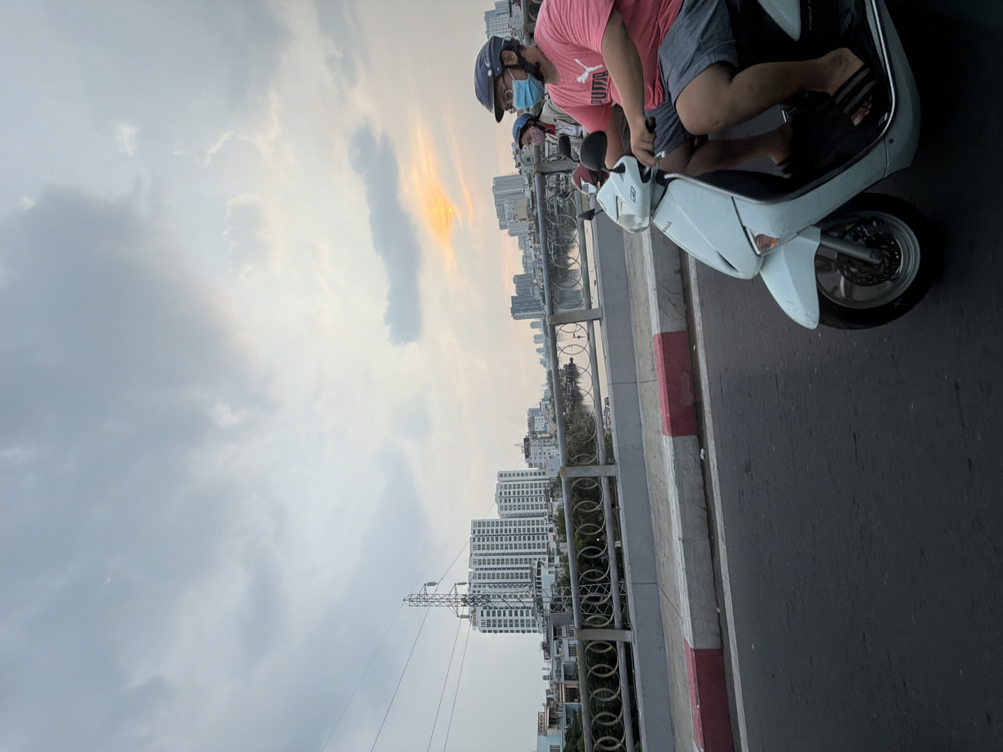 This vertical, outdoor, medium-distance shot shows a man riding a light blue scooter on a road in Thủ Đức City, Vietnam, on an overcast day with the sun breaking through the clouds in the distance. The man, wearing a blue helmet, a light blue face mask, a pink t-shirt, and gray shorts, is driving the scooter in the foreground and facing to the right. Behind him, a second scooter, partially obscured, carries a passenger wearing a pink and white mask and a light-colored jacket. The scooter is on a bridge with a grey and red striped curb. In the background, a body of water is visible, reflecting the overcast sky and the city skyline, which includes several tall, modern buildings and a large electrical tower. The overall impression is one of urban activity against a backdrop of a cloudy sky with patches of sunlight.