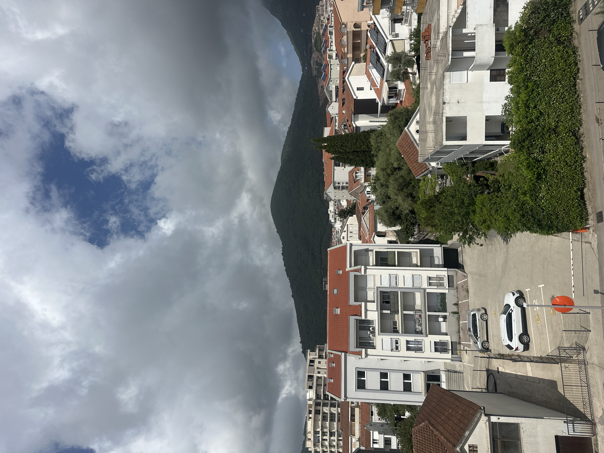 The image shows a view of Budva, Montenegro, from an elevated position. A mountain partially obscured by heavy clouds dominates the left side of the frame. The right side depicts a dense cluster of buildings, mostly white with red tile roofs, typical of the region's architecture. In the foreground, a parking area is visible with two parked cars, one white and one silver, along with parking space markings. A barrier pole with an orange circle is situated in the parking lot. Lush green vegetation is visible along the edges of the buildings and road, creating a contrast with the urban environment. The sky above is a mix of heavy clouds and patches of blue.