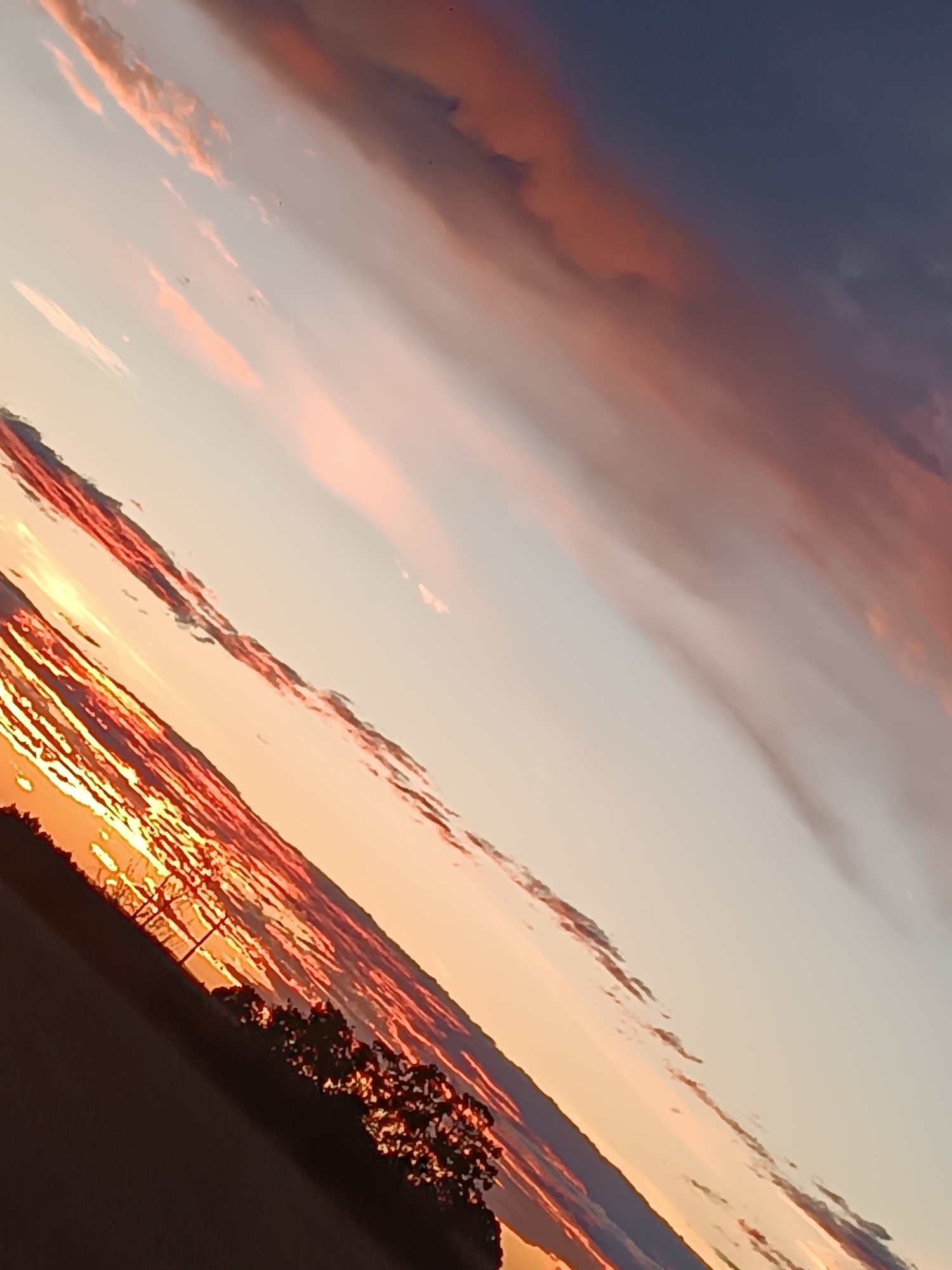 The image displays a diagonal composition of a sunset over a rural landscape, likely outside of Viadana, Italy. The sky is filled with dramatic clouds, transitioning from a deep indigo at the top to hues of orange and pink closer to the horizon. The sun, or its direct rays, is visible at the lower left, illuminating thin, streaky clouds with intense orange and red light. Silhouetted against the vibrant sky are trees and a pole, possibly a utility pole, suggesting a sparsely populated area. The lower portion of the image is a dark, indistinct ground or field. The overall impression is one of a vivid and atmospheric sunset.
