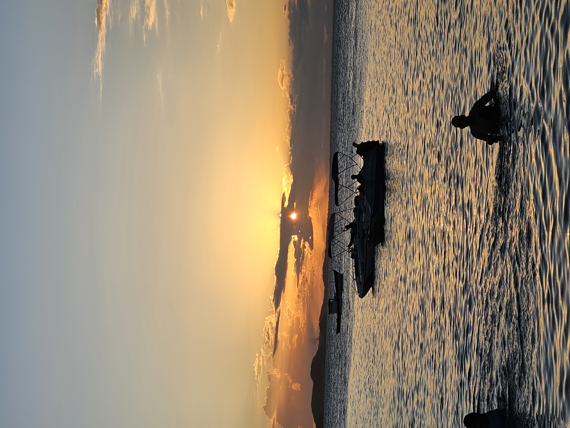 This photo captures a sunset scene on the water near Armação Dos Búzios, Brazil. The sun is obscured by clouds, casting a golden glow across the sky and water. Silhouetted against the light are several boats and the figure of a person in the water. The water's surface is rippled, reflecting the sunset.