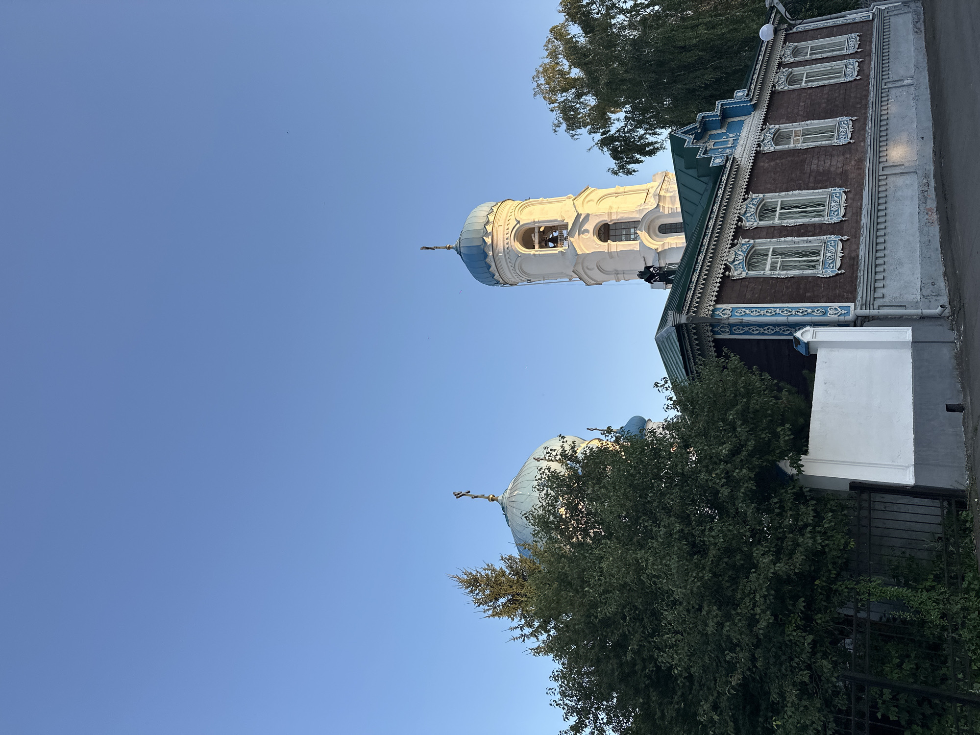 The image depicts a serene street scene in Biysk, Russia, under a clear blue sky. In the foreground, a traditional wooden house stands prominently, its dark brown facade adorned with intricate blue and white carved wooden trim around the windows and eaves, a hallmark of Russian folk architecture. The house has a green pitched roof and multiple windows with decorative grilles.
Behind the house, a white bell tower with a blue dome rises into the sky, topped with a cross. To the left of the bell tower, another smaller blue dome of a church is partially visible, also topped with a cross. A large, leafy green tree obscures part of the church dome and a portion of the bell tower. To the right of the bell tower, the branches of a tall, thin tree with sparse leaves are visible. A section of a white wall with a grey base is visible to the left of the house, adjacent to a dark metal fence covered in green foliage. The ground in front of the house is a paved road. A person, dressed in dark clothing and possibly wearing a helmet, can be seen standing on a ladder attached to the bell tower. The overall impression is of a peaceful historical district.