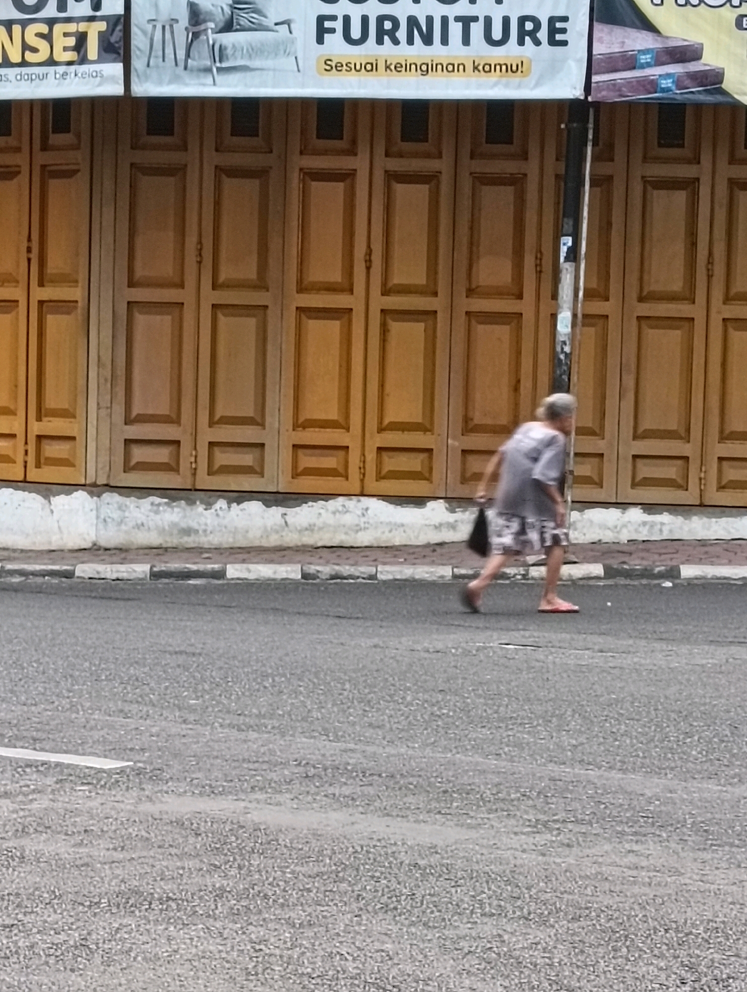 A slightly blurry, full-shot photograph depicts an elderly person walking on a street in Purworejo, Indonesia. The person, wearing a grey t-shirt and patterned shorts, is moving from left to right across the frame, carrying a dark bag in their left hand. They are wearing red flip-flops. The ground is an asphalt road with a white dashed line on the left side of the frame. To the right of the road is a sidewalk made of light-colored stones, bordered by a raised concrete curb. Behind the sidewalk is a building with large wooden doors that appear to be folded or paneled. Above the doors, a banner displaying text is partially visible, with "FURNITURE" being the most legible word. The overall lighting suggests it is daytime.
