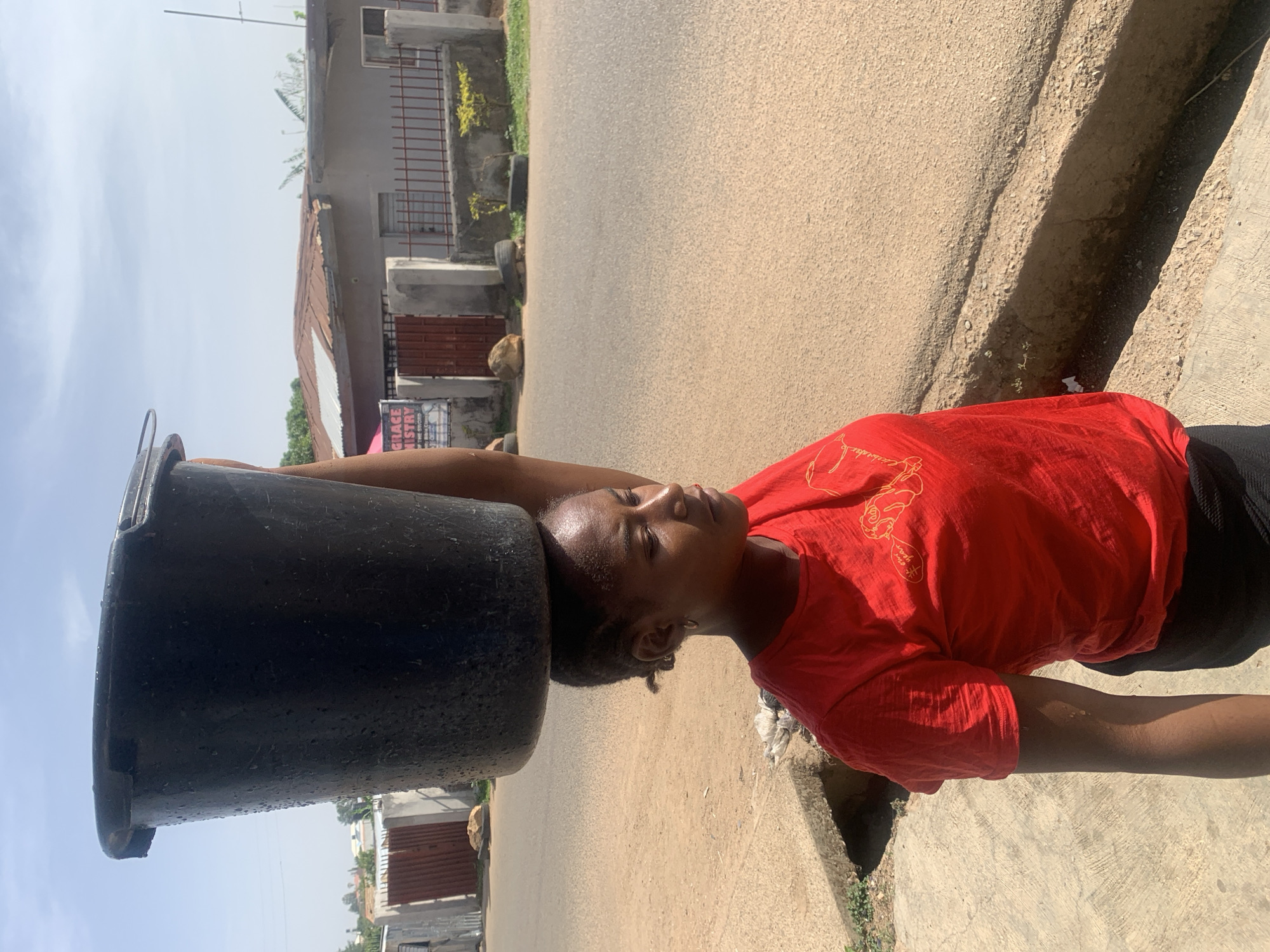An outdoor image shows a person balancing a black bucket on their head while standing on the side of a road in Jos, Nigeria. The person is wearing a red t-shirt and black bottoms. The road is paved and has a drainage ditch on the side where the person is standing. A building with a fence is in the background. The image is rotated 90 degrees counter-clockwise.