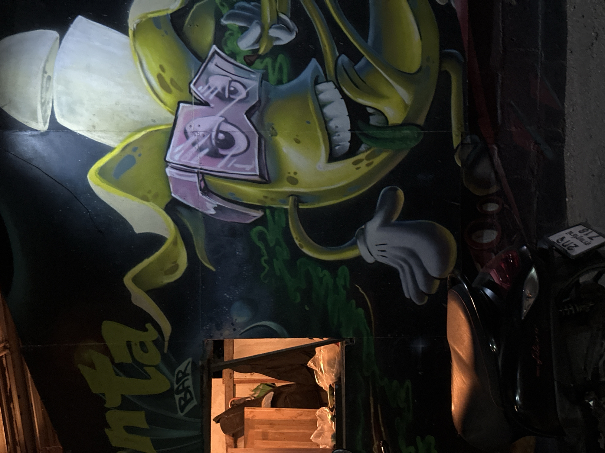 The image depicts a section of an exterior wall in ตำบลเกาะพะงัน, Thailand. The wall is covered with graffiti art featuring a yellow cartoon character, partially obscured, with white cartoon hands and a face overlaid with a pink "3" symbol. Lower on the wall, stylized text spells "NTA BAR" in green. A small, framed opening reveals a glimpse of an interior space with wooden structures and storage. Miscellaneous objects and a partially visible motorcycle with a license plate are situated near the right edge of the frame. The scene is lit dimly.