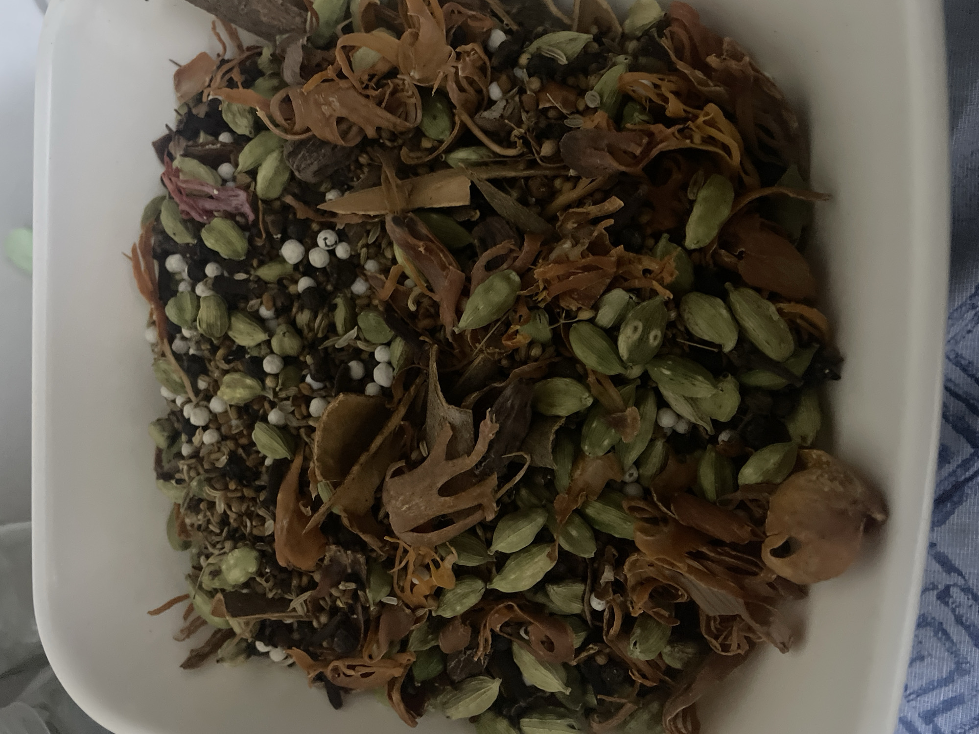 A top-down shot shows a white bowl filled with a mixture of spices. The most prominent spices include green cardamom pods, mace (the orange-brown, lacy covering of the nutmeg), and small white peppercorns. Other, smaller fragments of spices and seeds are interspersed within the mixture. The bowl is resting on a surface with a slightly blurred, blue patterned fabric. There is no specific information in the image that indicates it was taken in Dhaka, Bangladesh. There are no people, actions, or interactions depicted in the image.