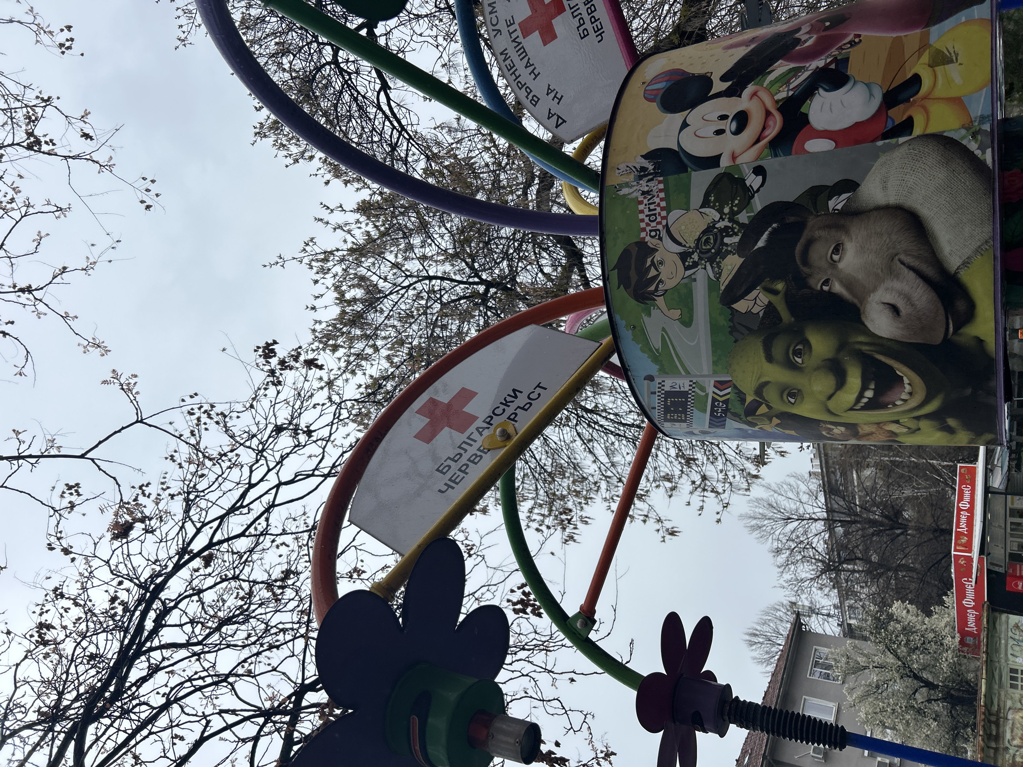 The image shows an upward angle shot of a playground structure in Plovdiv, Bulgaria, with cartoon images like Mickey Mouse, Shrek, and Ben 10. The structure also features signs, one with a red cross and Bulgarian text, and another with "Döner Finec" written in Bulgarian. The background includes bare winter trees, buildings, and a cloudy sky. The location appears to be urban, given the presence of buildings and signs.