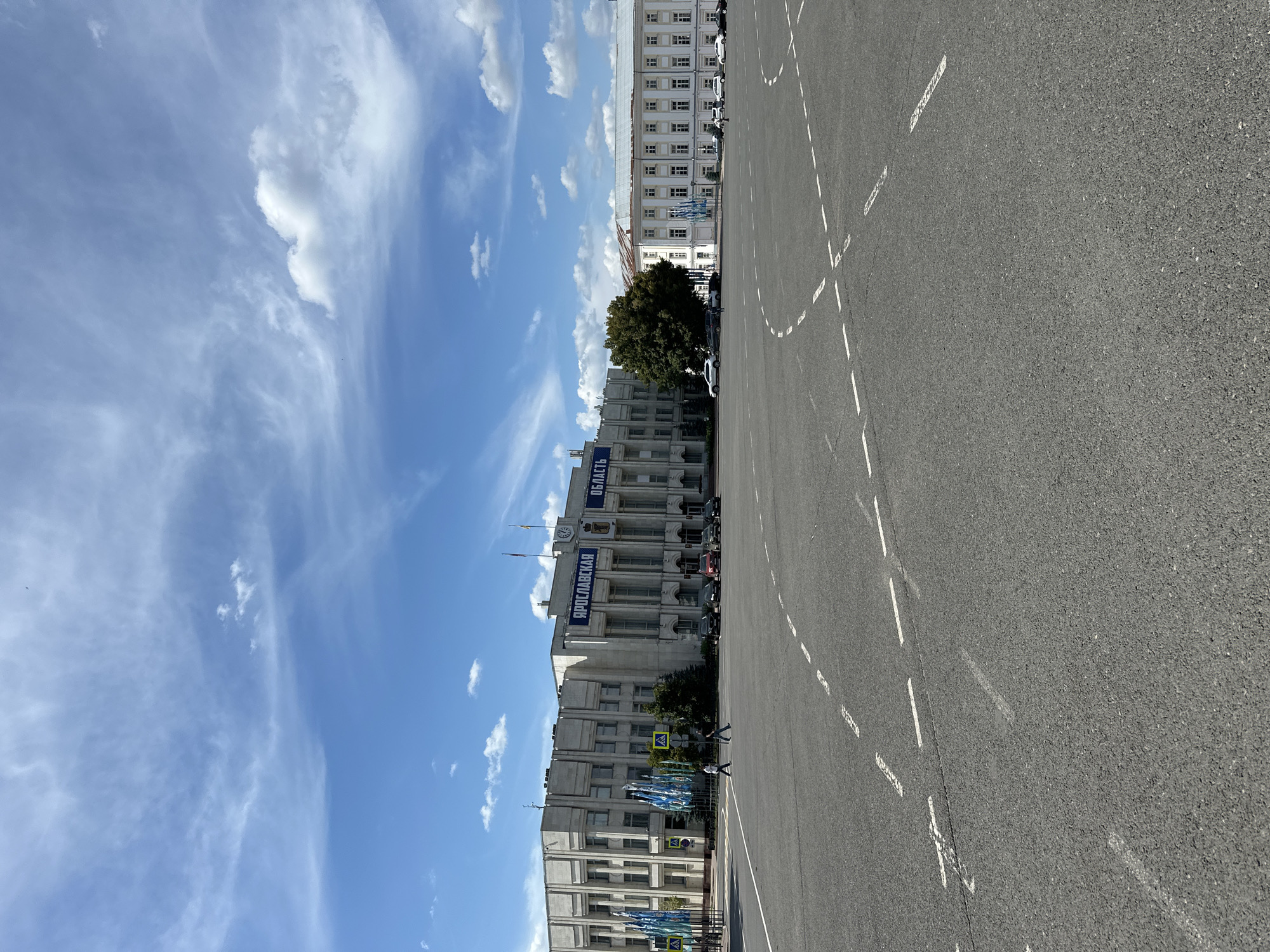 The image depicts a wide, paved public square in front of a large, imposing building with a neoclassical architectural style. The building is light-colored and features numerous windows, columns, and decorative elements. A large sign with Cyrillic lettering, reading "ЯРОСЛАВСКАЯ ОБЛАСТЬ" (Yaroslavskaya Oblast), is prominently displayed on the facade. Flags are visible on the building's roof, and a clock is mounted above the entrance.
To the left of the main building, a smaller structure with a similar architectural style is visible, adorned with several tall, blue banners. Trees and greenery are situated between the two buildings and on the far side of the main building. Several cars are parked in front of the main building and along the side of the square. Two individuals are walking across the paved area in the foreground. The square itself is marked with dashed white lines, indicating traffic lanes or parking spaces. The sky above is a bright blue with scattered white clouds, suggesting fair weather.
While the image captures a scene in Russia and features the text "Yaroslavskaya Oblast" (Yaroslavl Oblast), the specific location of Putyatino (Semivragovskaya S/A) cannot be definitively confirmed or denied based solely on the visual information provided in the image. Yaroslavl Oblast is a federal subject of Russia, and Putyatino could be a town or village within this region. However, the building appears to be a significant administrative or governmental structure, more likely to be located in a larger city or regional center within Yaroslavl Oblast rather than a small village named Putyatino.