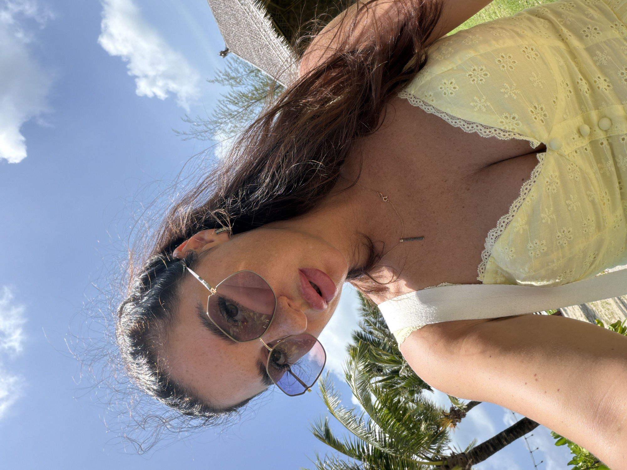 A woman stands outdoors on a sunny day, her face angled towards the camera. She wears oversized, hexagonal sunglasses with pink lenses and a yellow, eyelet-fabric camisole with white lace trim and a button-front closure. A delicate gold necklace with a small pendant is visible on her chest. Her dark brown hair is windswept, with strands framing her face and falling over her shoulders. In the background, palm trees are visible, and a thatched-roof building stands to the right. The sky is bright blue with scattered white clouds. The location is identified as Bang Muang, Thailand.