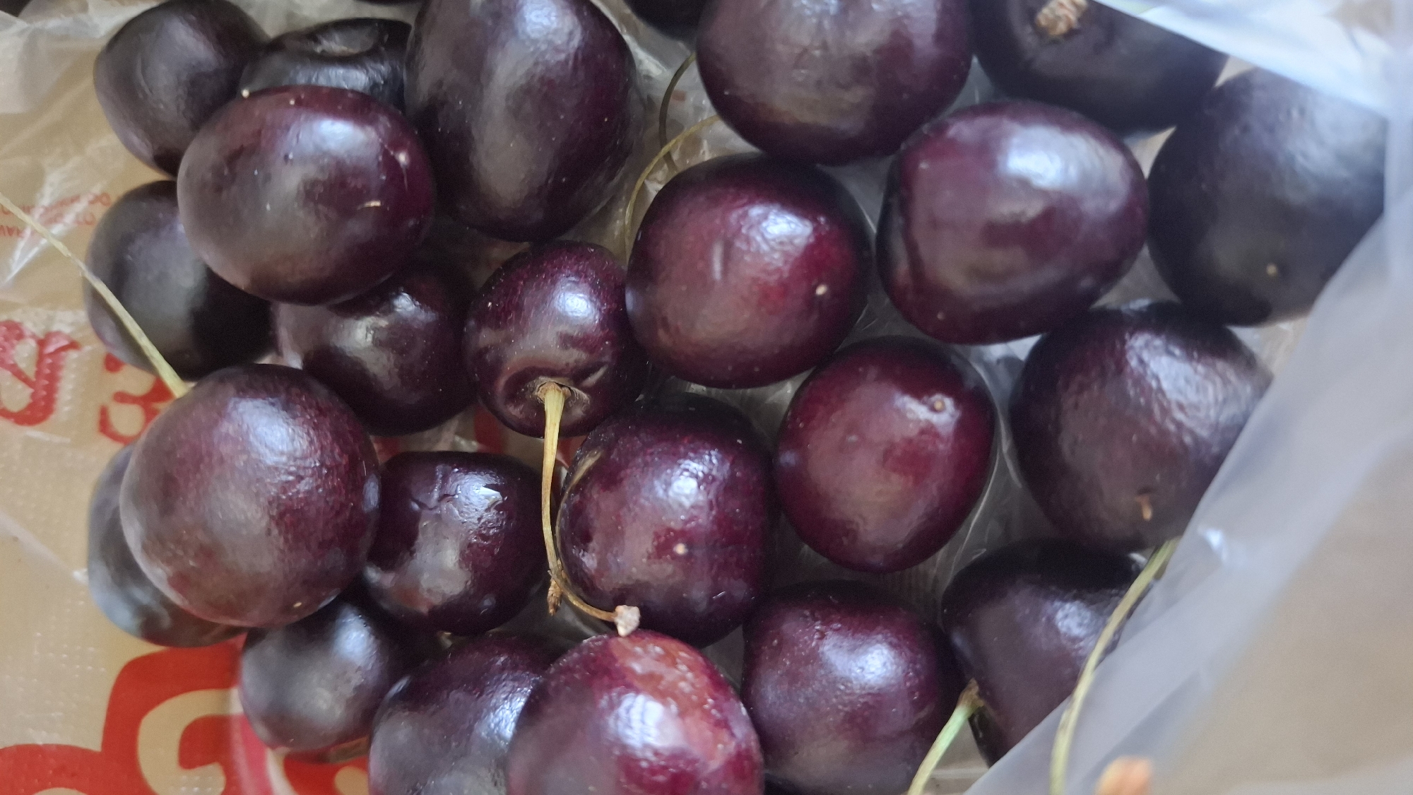 A close-up, overhead shot shows a pile of ripe, dark red cherries inside a clear plastic bag. The cherries are round and glossy, with some stems still attached. The bag is partially opened, revealing a glimpse of a red and white pattern on what appears to be a paper shopping bag underneath. The background is blurred and out of focus. The scene is indoors, with natural light illuminating the fruit. There are no people present. The image does not contain any elements that would indicate a specific location in Shymkent, Kazakhstan.