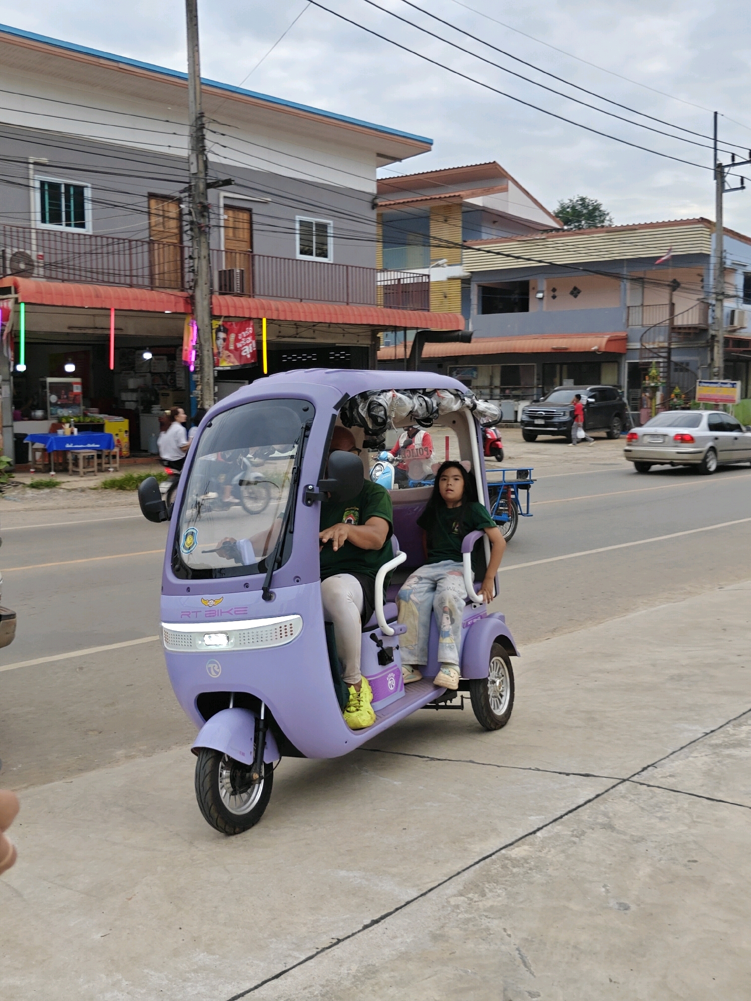 In Mu Si, Thailand, a person is driving a lavender three-wheeled electric scooter on a paved road. The scooter has a canopy overhead. A young girl with long dark hair is seated next to the driver. The girl is wearing a dark green t-shirt and light blue pants with a Disney princess pattern. She is looking towards the camera. The driver is wearing a dark green t-shirt and light-colored pants, with a black face covering. Yellow shoes are visible on the driver's feet. In the background, there are buildings with balconies and awnings. A silver sedan is driving on the road, and other vehicles and a person on a motorcycle are visible further in the distance. The sky is overcast.