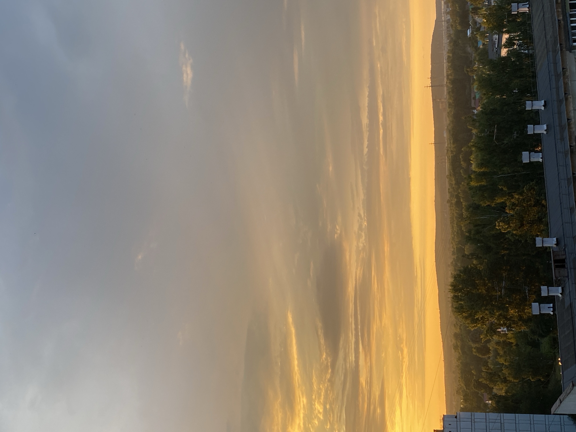 The image depicts a sunset over the city of Chaikovsky, Russia. The sky is filled with dramatic clouds, transitioning from a bright orange and yellow near the horizon to a muted grey and blue overhead. The sun is low, casting a warm glow on the landscape below. A dense line of green trees stretches across the middle ground, interspersed with some buildings. In the foreground, the roof of a low-rise industrial building is visible, characterized by its dark, flat roof and a series of white rectangular chimneys or ventilation shafts. To the far left, a portion of a multi-story apartment building, with a light-colored facade and visible window panes, is also present. In the distance, hills and power lines are faintly discernible against the horizon. There are no people or animals visible in the frame. The overall scene suggests an urban fringe environment at the end of the day.
