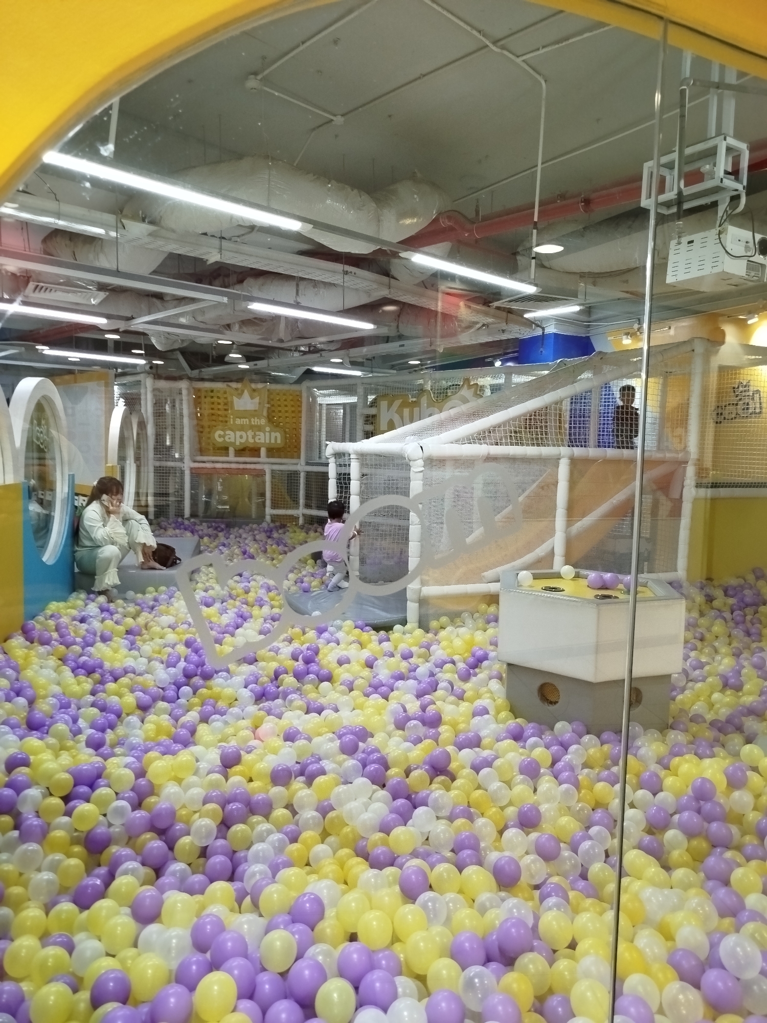 The image shows an indoor playground filled with a ball pit, seemingly within a commercial establishment. The ball pit contains yellow, white, and purple balls. A woman, wearing a mask, is seated at the edge of the pit, apparently talking on a mobile phone. A young girl is standing near the climbing structure with netting, another person is at the top of the climbing structure. Behind them, a sign reads "I am the captain". The playground equipment includes slides and tunnels. The environment appears to be well-lit with overhead fluorescent lights, suggesting a retail or entertainment space. The location is possibly Da Lat, Vietnam, given the 'Kubilat' sign on the wall behind. There are ceiling fixtures and ventilation systems visible above the play area.
