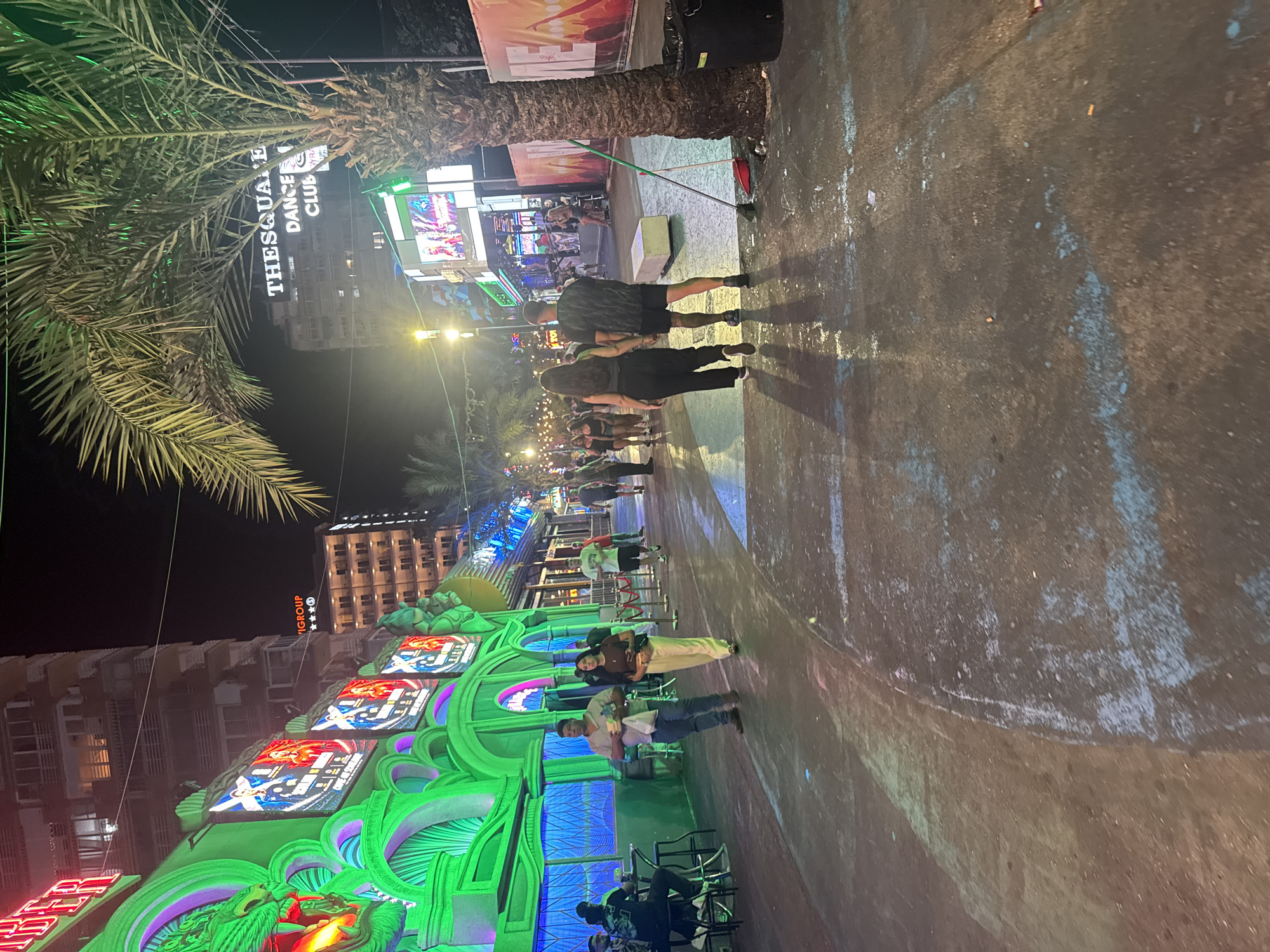 The image depicts a street scene at night in Benidorm, Spain, likely on or near the famous "Street of the Ripper" (Calle Gerona). The area is characterized by bright neon lighting from various entertainment venues and palm trees lining the walkway.
In the foreground, a paved street or promenade is visible, with patches of blue and white paint or chalk marks. Several people are walking along the street. Two individuals, a man and a woman, are walking away from the camera towards the right. The man wears a dark t-shirt and shorts, while the woman is in dark pants and a dark top. Further down the street, a couple also walks away from the camera, the man in a dark patterned t-shirt and shorts, and the woman in black pants and a dark top. Other individuals are scattered throughout the scene, some walking, some standing or sitting at outdoor seating areas.
On the left side of the image, a building with vibrant green neon lights and illuminated screens displaying images of people can be seen. The screens appear to advertise entertainment or events. Above this, a multi-story building with numerous windows is visible, and further in the background, another tall building with the text "THE SQUARE DANCE CLUB" illuminated in white is prominent. The overall atmosphere suggests a lively nightlife district.
To the right of the frame, a tall palm tree stands near a black trash bin and a red-handled broom and dustpan, suggesting some recent cleaning or maintenance. The ground is concrete, and there are shadows cast by the people and the palm trees, indicating the presence of artificial lighting. The image captures a typical nighttime scene in a tourist-heavy entertainment area.