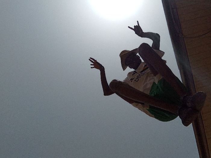 A single individual is depicted outdoors in Narayi, Nigeria, captured from a low-angle perspective against a bright, hazy sky with the sun visible in the upper right. The subject, wearing a light-colored straw hat, a white t-shirt with 'betss' branding, green shorts, and dark shoes, is positioned diagonally in the frame. Their right arm is raised, with the hand performing a 'rock on' gesture (index and pinky fingers extended). The left hand is also raised, with fingers partially splayed. The person appears to be leaning against or supported by a vertical wooden or metal post visible on the right side of the frame.