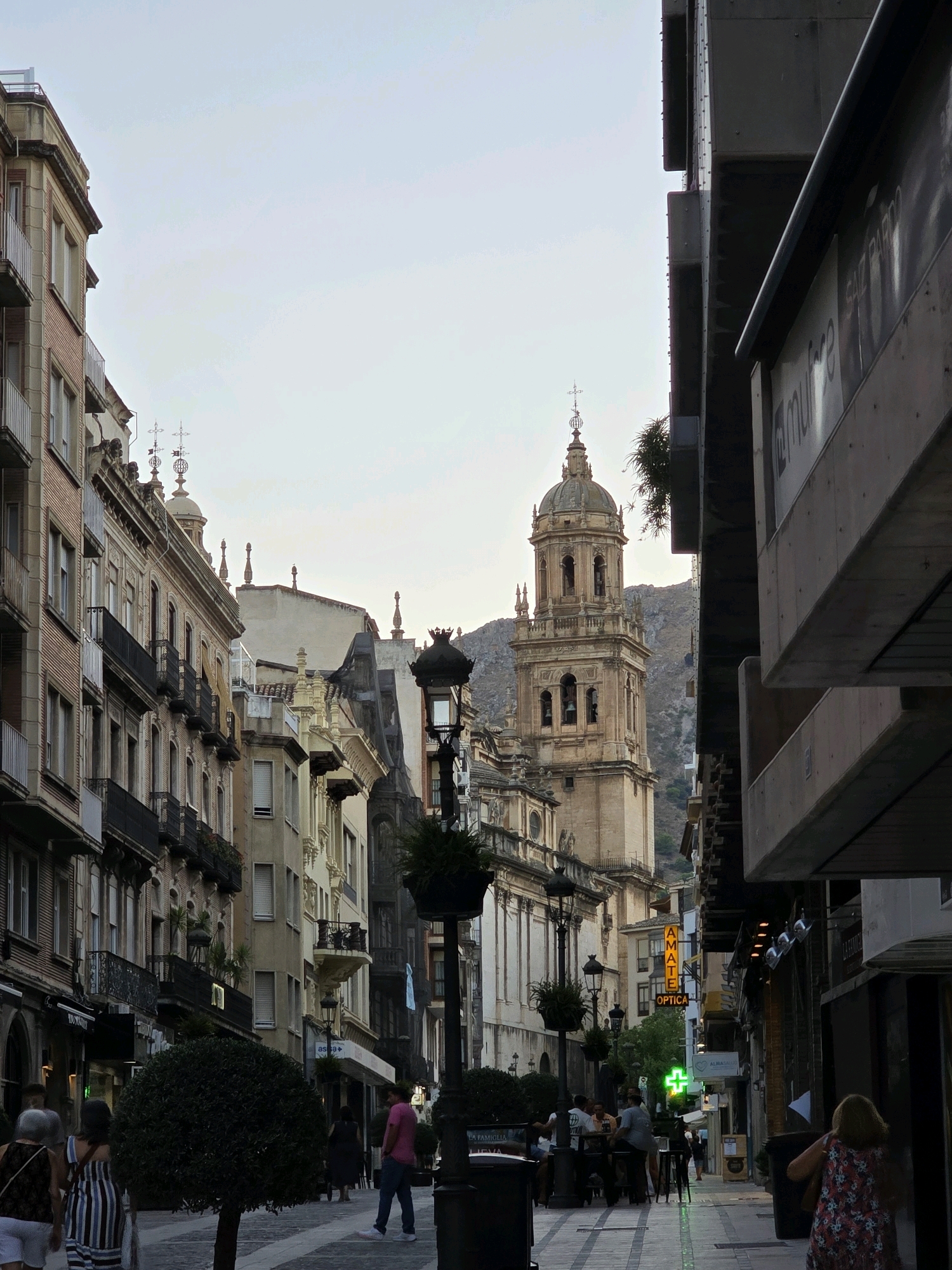 The image depicts a street scene in a city, likely in Europe, with a mix of old and new architecture. A prominent bell tower of a cathedral or church dominates the background, its classical design contrasting with the modern buildings lining the street on the right. The street is paved with cobblestones and lined with shops, cafes, and restaurants.
Several people are visible in the scene. On the left side of the street, a group of people are seated at tables outside a cafe. Further down the street, a man in a pink shirt and jeans walks towards the viewer, while a woman in a floral dress walks away from the viewer. On the right side of the street, a person is seen entering a building.
The atmosphere of the scene is one of a typical urban environment, with people going about their daily activities. The lighting suggests it is late afternoon or early evening, with the sky a pale blue. The presence of a green cross sign indicates a pharmacy.