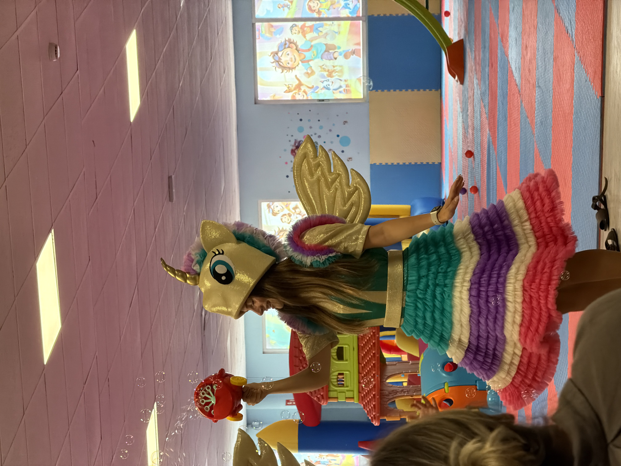 The image shows a woman dressed as a unicorn, holding a red bubble machine and blowing bubbles. She is wearing a gold unicorn hat with a rainbow mane, gold wings, and a multi-colored ruffled skirt in teal, white, purple, and pink. The background appears to be a children's play area with brightly colored walls, soft flooring, and climbing structures. There are several bubbles visible in the air, and red balls are scattered on the floor. In the foreground, the back of a person's head and shoulders are visible, suggesting they are an observer. The scene is indoors, possibly a daycare, entertainment center, or party venue.