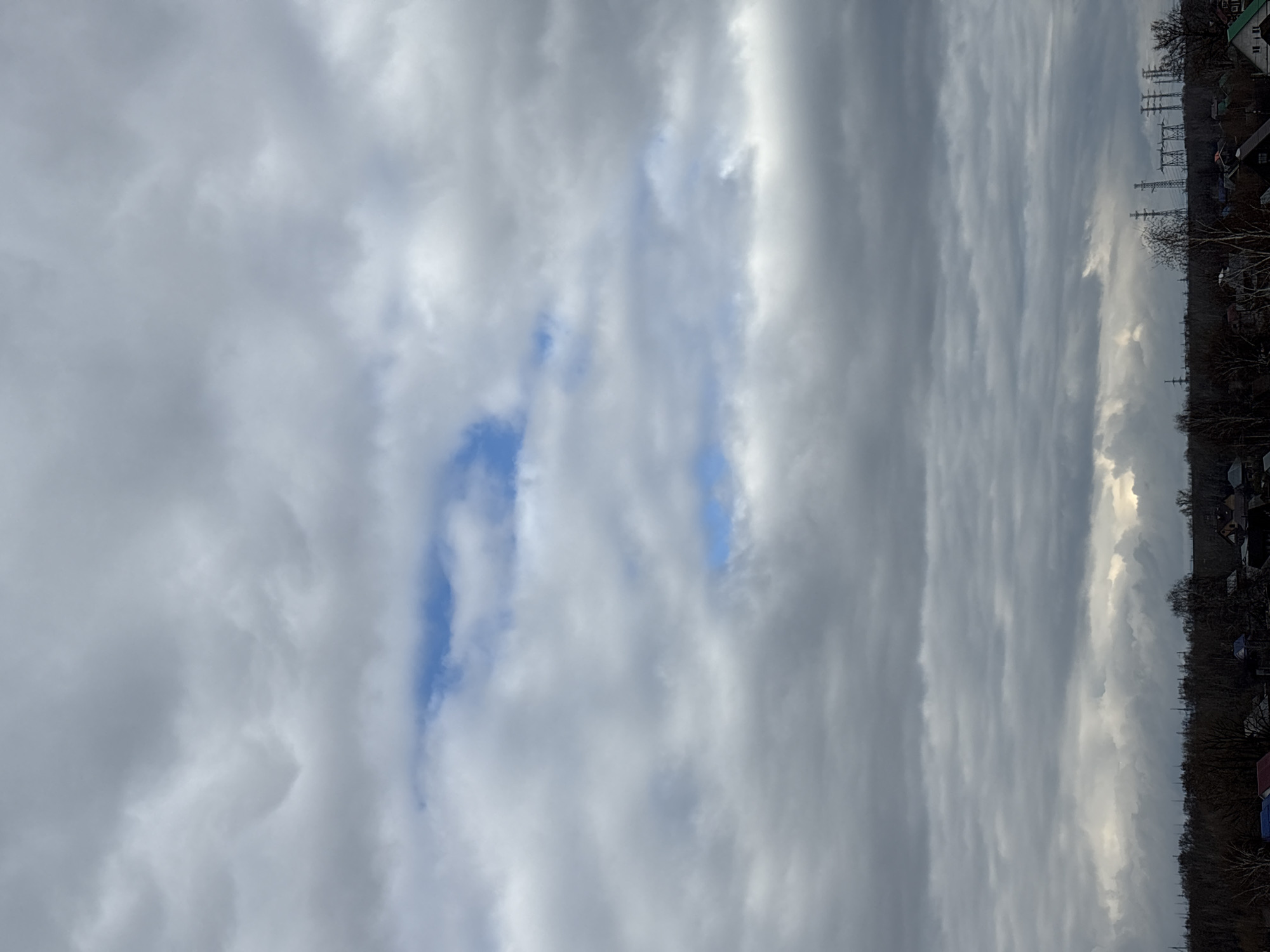 The image displays a sky dominated by a large expanse of clouds, with a distant horizon line visible at the bottom (when viewed in correct orientation). The clouds are primarily shades of grey and white, creating a textured and dynamic overhead scene. Through breaks in the cloud cover, patches of bright blue sky are visible, particularly towards the center of the frame, suggesting a partially cloudy day rather than a completely overcast one. Some of the white clouds appear brighter, possibly catching sunlight.
Along the distant horizon, a dark, dense line of trees or forest stretches across the landscape. Interspersed within and above the tree line, particularly on the right side of the image, are several tall utility poles with power lines, indicating electrical infrastructure. A few dark-roofed buildings or houses are also faintly visible nestled within or behind the tree line.
The scene depicts an outdoor environment, likely in the daytime given the visible light, though the heavy cloud cover suggests it might be an overcast day or perhaps late afternoon. There is no visible activity or specific event taking place; it is a static landscape view. The setting, with its blend of natural elements like trees and clouds alongside man-made structures like power lines and buildings, suggests a semi-rural or suburban area. Based on the provided context, the location is Chaikovsky, Russia. There is no visible text in the image.