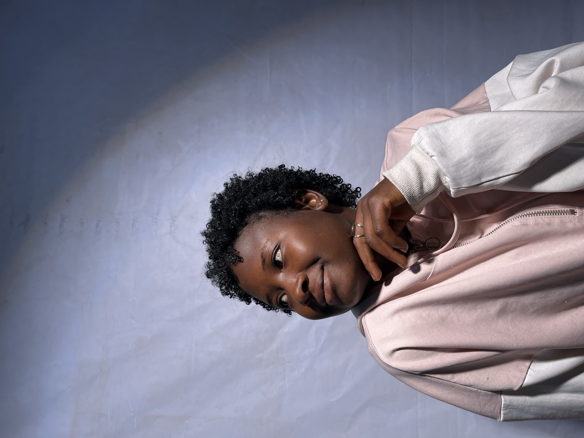 A young Black woman with short, curly dark hair and dark skin is captured in a medium shot against a textured, light purple backdrop. She is looking off to her left with a slight smile, her chin resting on her left hand, which is adorned with two rings. She is wearing a pink and white color-blocked hooded jacket. A diagonal band of light and shadow cuts across the upper right portion of the background, suggesting artificial lighting or a window. The woman's pose and expression are relaxed and contemplative. There are no discernible elements in the image that would allow for the specific identification of her location within the city of Jos, Nigeria.