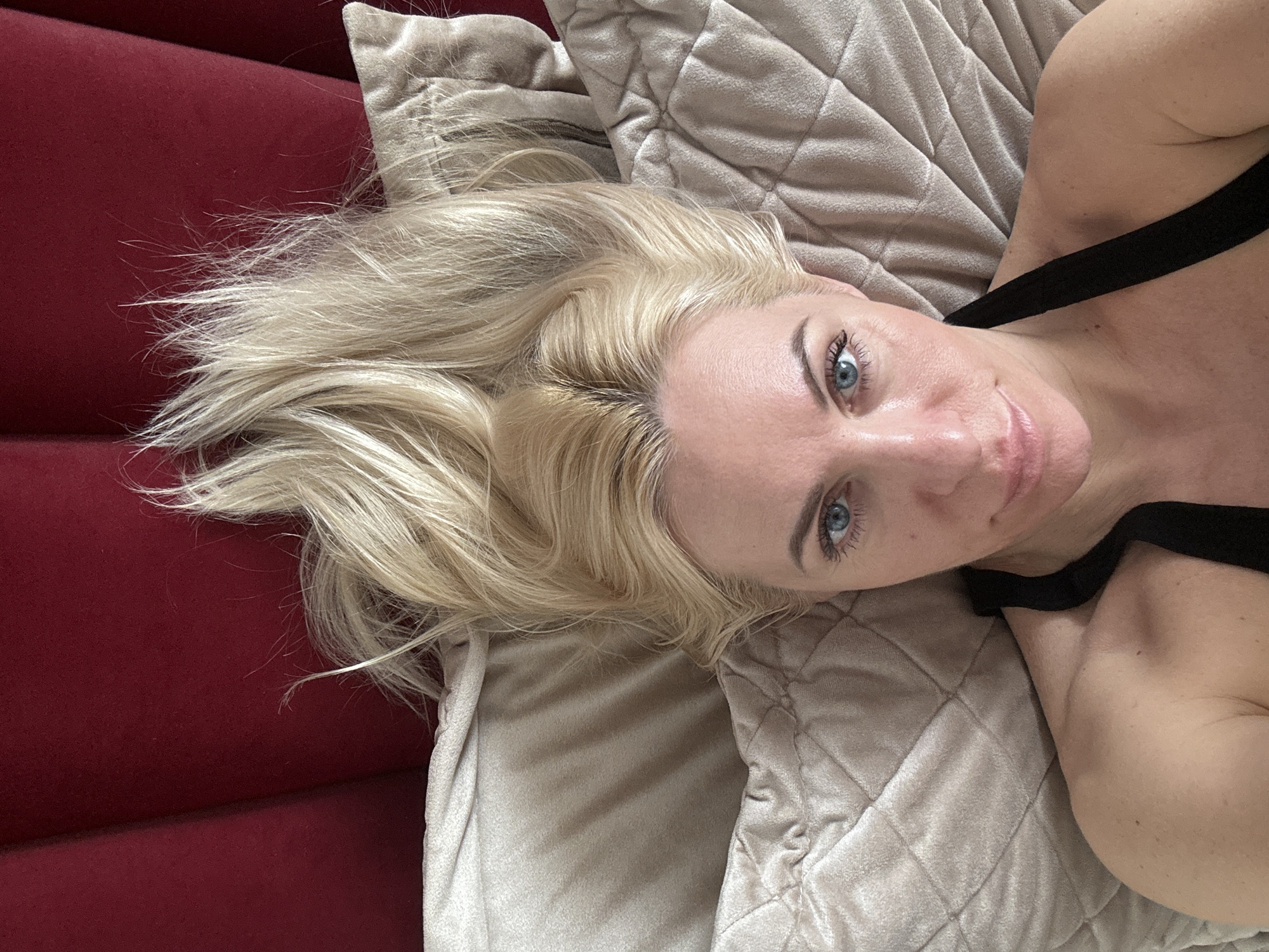A blonde-haired woman with blue eyes is lying on a beige quilted pillow, next to a red upholstered surface. She is wearing a black strap that crosses her chest. The location is Unknown, Unknown.
