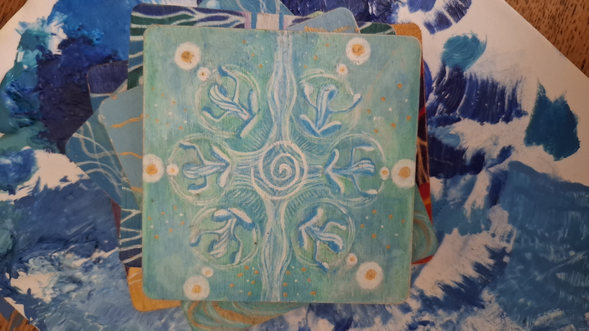 The image displays a centrally positioned, square-shaped, hand-painted panel or tile with slightly rounded corners. The panel features a light teal background adorned with a symmetrical, stylized design in white and light blue. At its core is a white spiral, from which an elongated, undulating white form extends vertically. Surrounding the central spiral are eight stylized, curvilinear leaf-like or floral motifs rendered in white and shades of blue, arranged in a circular pattern, with some enclosed within white circular outlines. The teal background is dotted with numerous small orange or gold flecks, and four larger white circles with orange centers are positioned near the cardinal points of the design.
This main panel rests upon a layered arrangement of other painted materials, primarily sheets of paper or cardstock. These underlying elements are characterized by abstract, textured brushstrokes in various shades of blue (from dark navy to light sky blue) and white, suggesting a vibrant artistic palette or a collection of painted scraps. Some pieces show hints of gold or yellow lines. A small section of a light brown, possibly wooden, surface is visible at the bottom-left corner of the frame. There are no people, animals, or direct actions depicted. The scene contains no identifiable geographical markers to indicate a location in Rassvet, Russia.