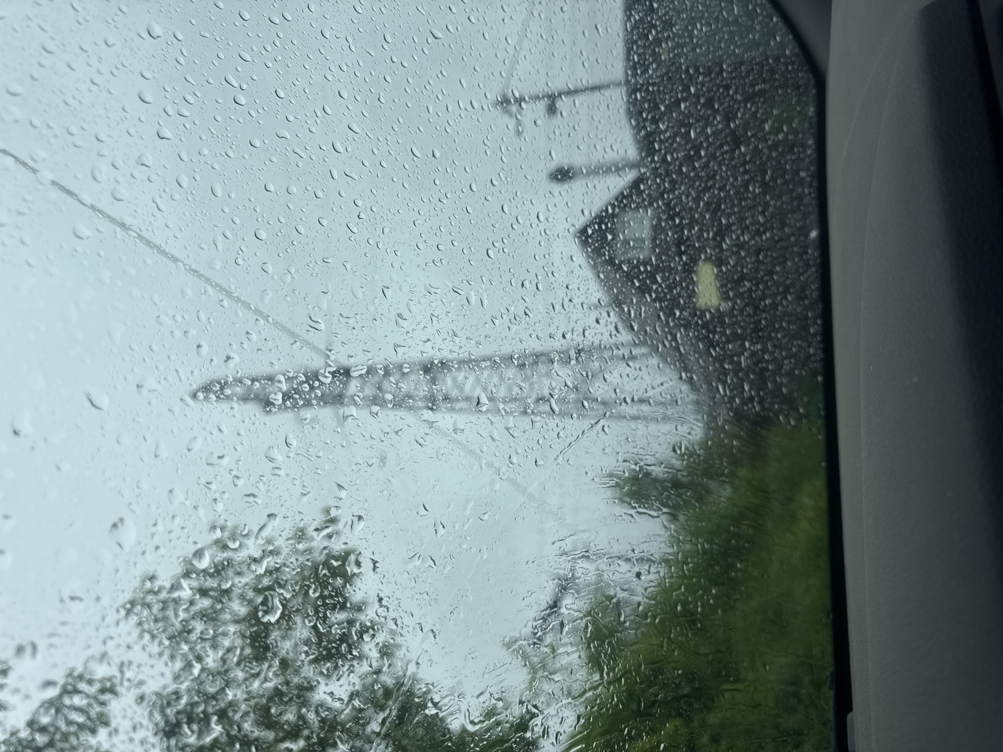 The image shows a view from inside a vehicle through a rain-covered window. Outside, trees and what appears to be a bridge structure are visible through the blurred raindrops. A street light or electrical pole is also partially visible. The location is potentially Chaikovsky, Russia, based on contextual information (the source of the image). The overall scene depicts a rainy day. The inside of the vehicle is visible on the right side of the frame.