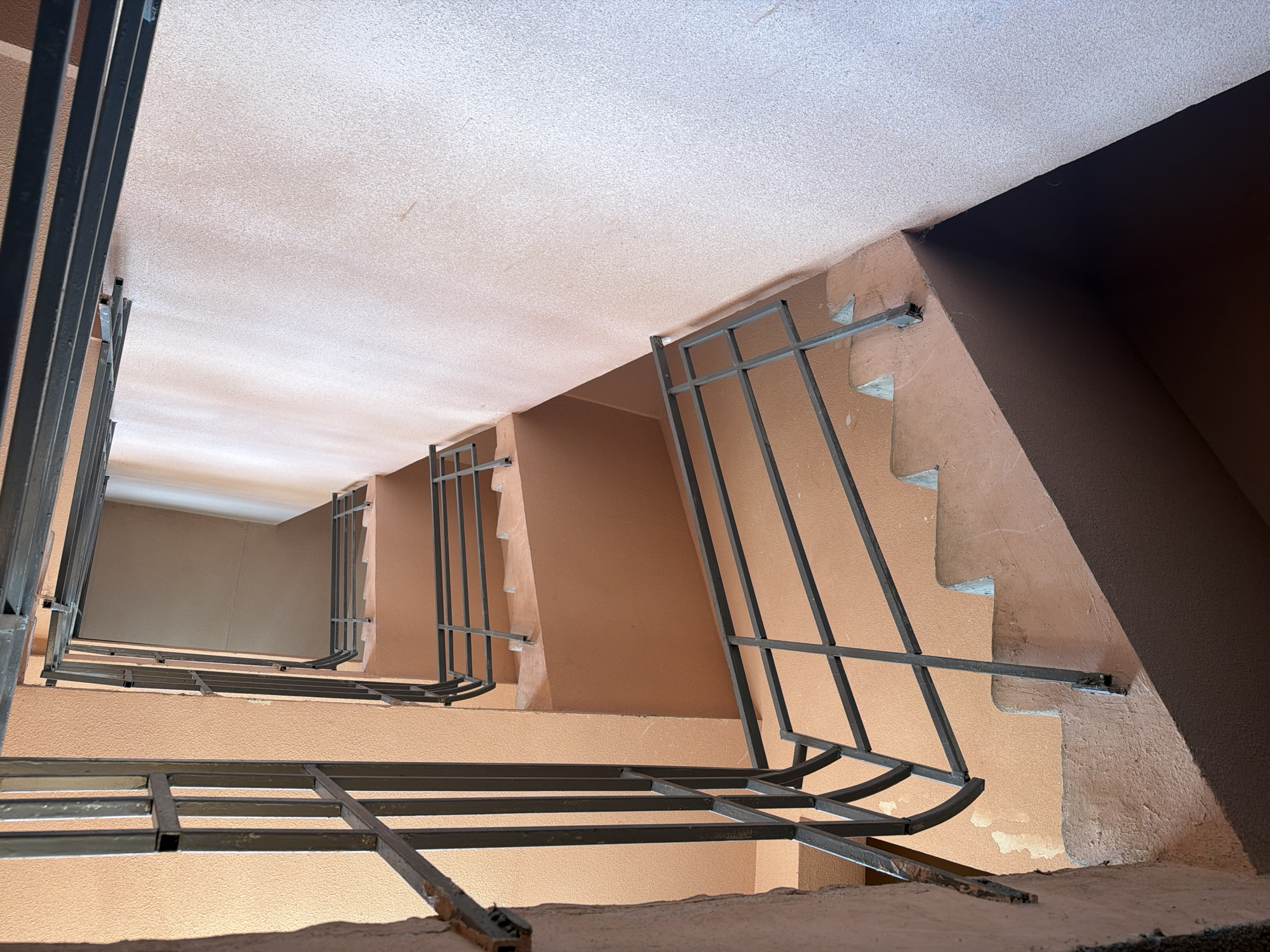The image shows the interior of a concrete stairwell, likely in an apartment building in Lebedyan, Russia. The walls are painted a light peach color, and the concrete stairs are also a similar hue, appearing worn and slightly damaged in places. Metal railings, painted dark gray, line the sides of the staircases. The perspective is from a low angle looking upwards, revealing multiple landings and flights of stairs ascending into the building's core. The lighting suggests an indoor environment with no direct sunlight. There are no people or specific objects that indicate a particular activity or event. The scene depicts a utilitarian and somewhat aged building interior.