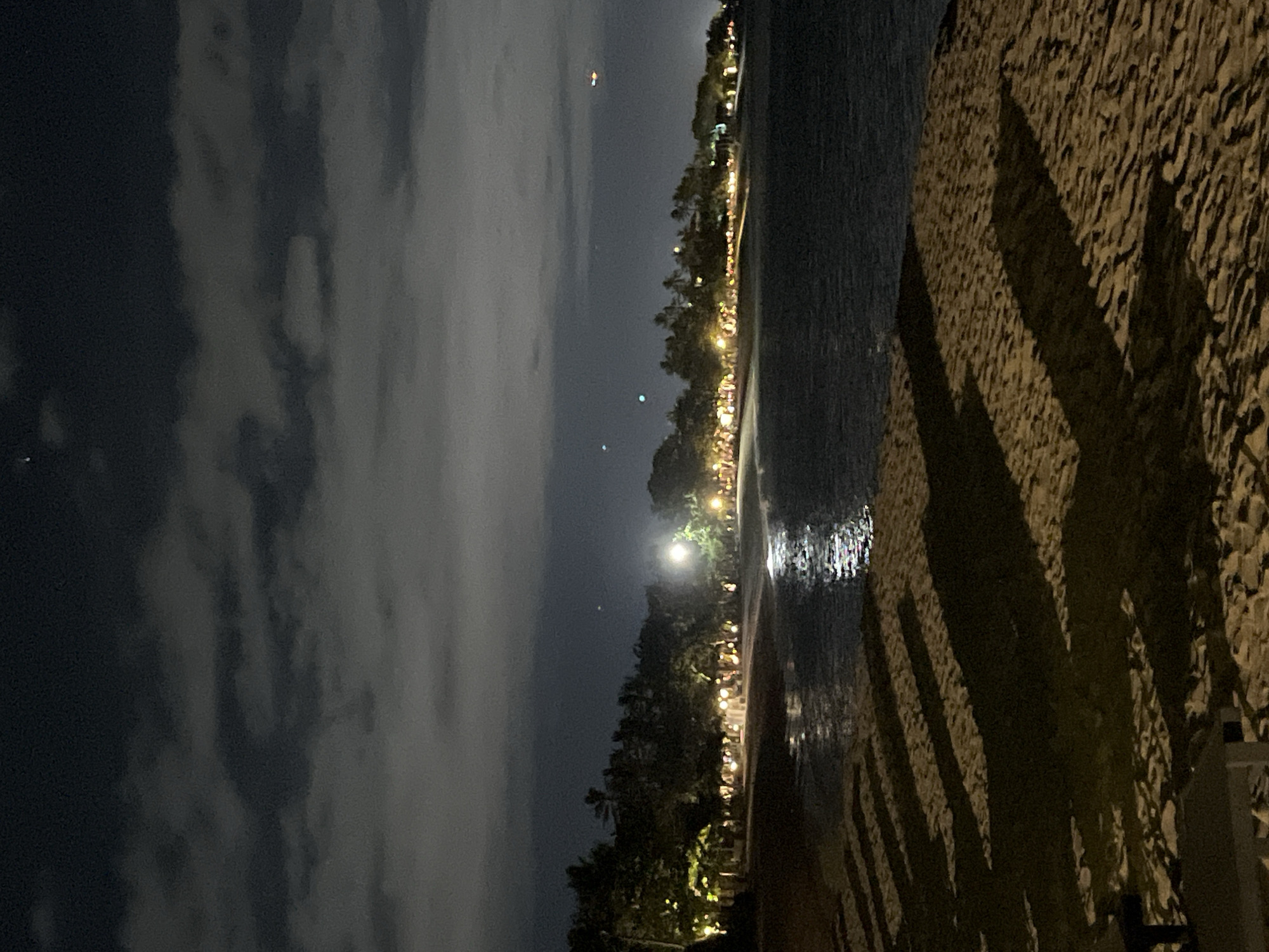 This is a nighttime shot of a beach. A body of water extends from the center of the frame towards the right. The immediate foreground contains an expanse of coarse sand with sharp shadows of what appear to be palm trees falling across it. Beyond the sand and bordering the water's edge, there is a line of trees with lights scattered throughout, indicating some form of development or resort. In the distance, more lights appear to stretch along the coastline. The sky is predominantly filled with clouds, though hints of moonlight are present, casting a faint glow. There is a single, bright star or light source visible above the trees. The location is Unknown, Indonesia.
