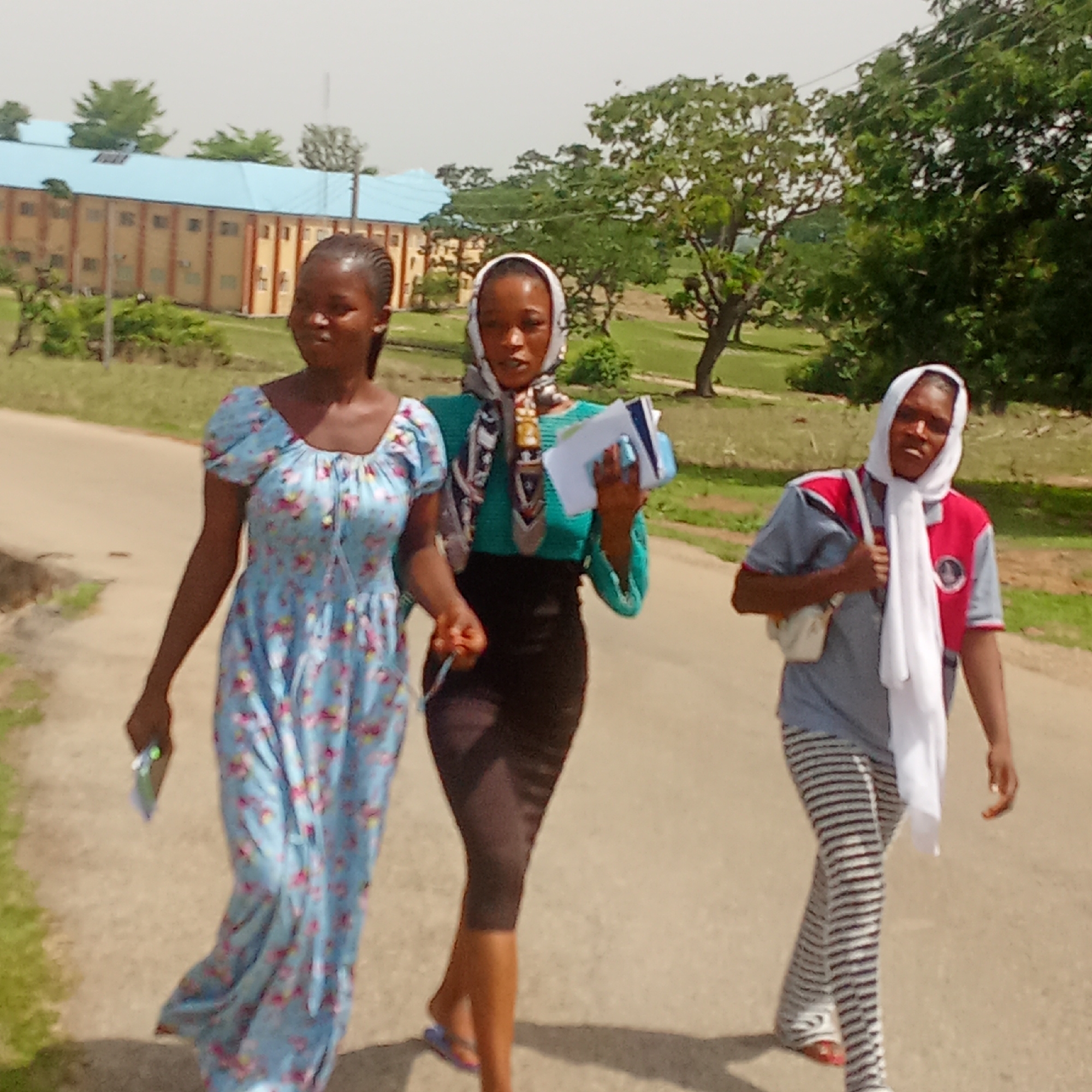 Three young women are captured walking along a paved road under bright daylight in Akwanga, Nigeria. The background features multi-story buildings with distinctive light blue roofs, indicative of a campus or institutional setting, surrounded by numerous green trees and grassy areas.
From left to right:
The first woman wears a light blue, long floral dress and has braided hair. She holds a small, rectangular object, possibly a phone, in her left hand.
The central woman is dressed in a teal long-sleeved top and a dark, fitted skirt, complemented by a patterned headscarf. She carries several papers or notebooks along with a pen, suggesting she might be a student or involved in an academic activity. She wears flip-flops.
The third woman on the right is attired in a grey and red short-sleeved collared shirt over horizontally striped pants. A white headscarf covers her head and neck, and she carries a light-colored shoulder bag. She also wears flip-flops, which appear red.
The women appear to be engaged in a casual stroll, possibly commuting to or from a destination within the campus or institutional grounds. The strong illumination and distinct shadows indicate it is likely midday or early afternoon. The scene conveys a sense of ordinary daily life. No discernible text is visible on any objects, clothing, or in the environment.