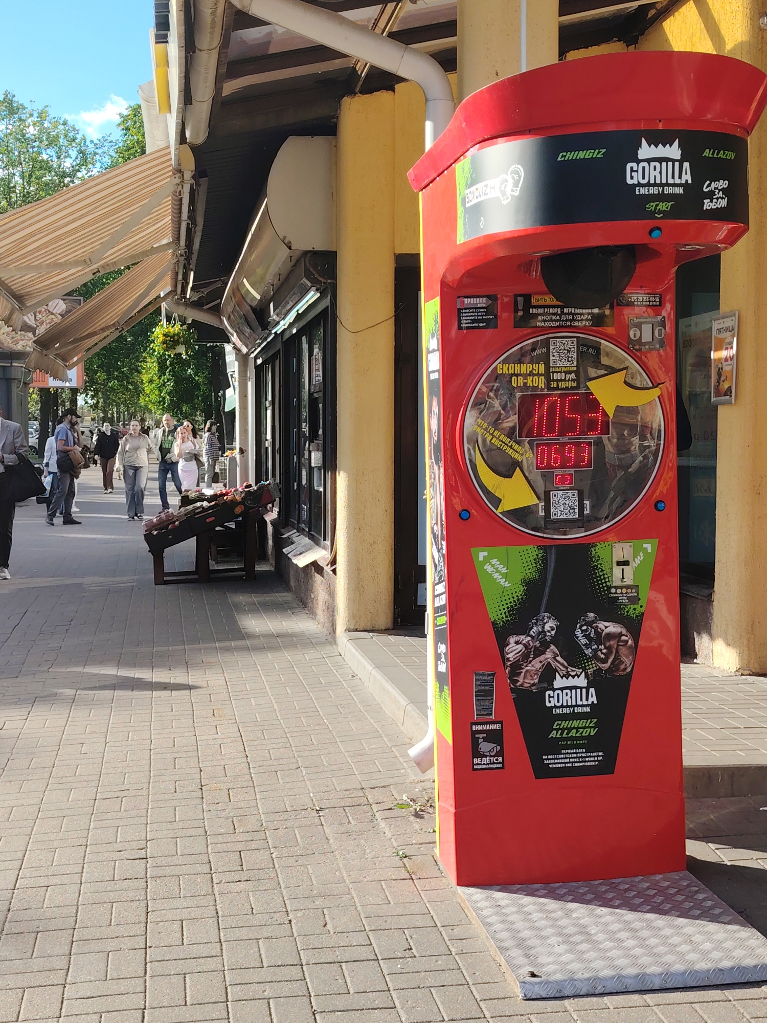 An outdoor scene in Minsk, Belarus, depicts a bustling city sidewalk on a sunny day. The primary subject is a large, red arcade punching machine, branded "GORILLA ENERGY DRINK" featuring the fighter "CHINGIZ ALLAZOV." The machine has a black top section with a punching bag and a digital display showing "1053" and "0693." Instructions in Russian are visible, including "КНОПКА ДЛЯ УДАРА НАХОДИТСЯ СВЕРХУ" (The button for the punch is at the top) and a prompt to "СКАНИРУЙ QR-КОД" (Scan QR code) for "1000 руб. за удары" (1000 rubles per punch). Below the display, images of male and female boxers are shown with "MAN WOMAN" labels. A "ВНИМАНИЕ! ВЕДЁТСЯ" (Attention! Surveillance in progress) sign is also present. The base of the machine rests on a metal checkered plate.
The machine is positioned along a tiled sidewalk, next to a building with a yellow facade and several storefronts. One storefront has a striped retractable awning extended, providing shade. Further down the sidewalk, an open vendor's display stand holds various items, possibly produce. Numerous pedestrians are seen walking in both directions, indicating a lively urban environment. Trees and a clear blue sky are visible in the background, suggesting pleasant weather, likely late morning or early afternoon due to the bright sunlight and shadows. A sign in the background reads "ПЯТНИЦА" (Friday).