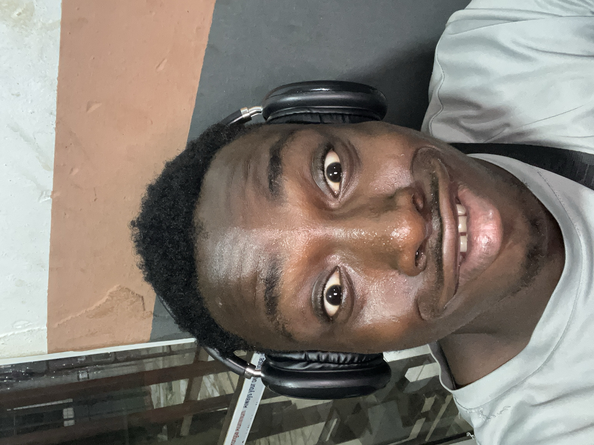 The image is a close-up, chest-up portrait of a dark-skinned person, appearing to be a man, looking directly at the camera with a wide smile. The original image is rotated 90 degrees counter-clockwise, showing the person lying on their right side, but for descriptive purposes, we'll assume an upright orientation.
The man has short, dark, curly hair and brown eyes. He is wearing black over-ear headphones, which rest on his head. His face, particularly his forehead, cheeks, and upper lip, appears noticeably sweaty, suggesting physical exertion or a warm environment. He is wearing a light grey, short-sleeved t-shirt. His expression is bright and engaged, with his mouth open in a smile that reveals his upper teeth.
The background is somewhat indistinct but appears to be an indoor or sheltered outdoor setting. To the man's left (our right, if upright), there's a section of a light brown or tan wall next to a lighter, possibly white or light grey, textured surface. To his right (our left), the background is a darker, possibly black or dark grey, surface. Below his chin, there's a reflective surface, likely a window or a metallic railing, which shows some structural elements in the reflection.
On a metallic bar visible in the reflective surface, some text is partially legible: "...ONGIAI ALUMINUM Always your de _oice". It likely reads "KONGIAI ALUMINUM Always your choice". The location provided is Nkpor, Nigeria, which aligns with a warm climate where one might sweat. The image captures a moment of direct engagement, possibly a selfie.