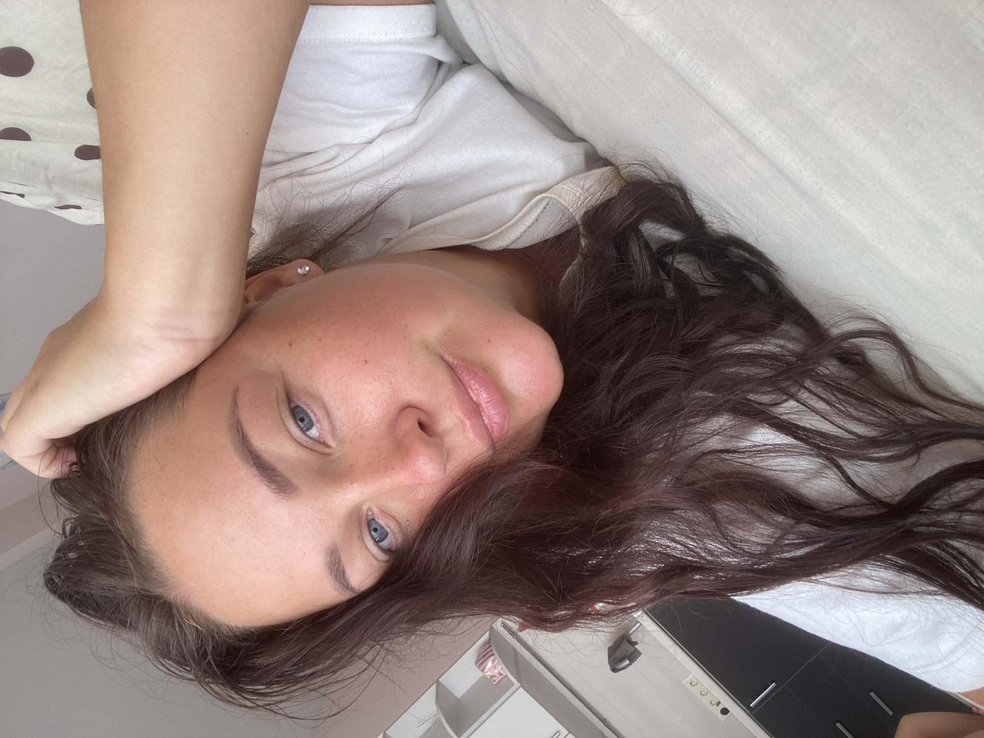 The image is a close-up, self-taken photograph of a woman lying down, possibly in bed. She has long, dark brown hair that is partially covering her face and shoulder. Her eyes are blue and open, and she is looking directly at the camera. Her left hand is resting on her head, and she is wearing a white t-shirt. The background shows a bed with white linens and a hint of a wall with some shelves and cabinets, suggesting an indoor setting like a bedroom or a hotel room. There is no indication of the city of Sochi, Russia.