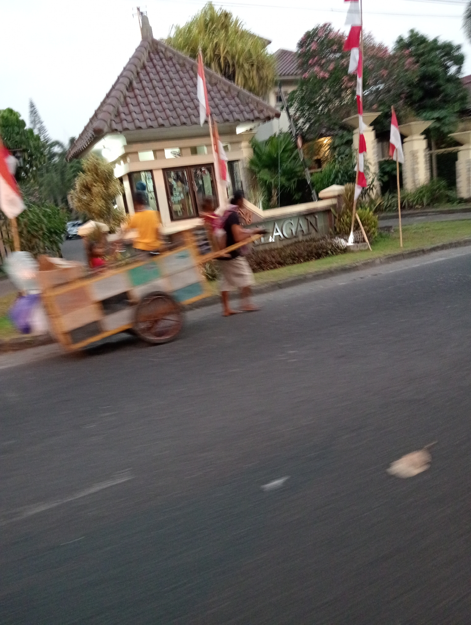 In Ngaglik, Indonesia, an out-of-focus scene depicts a street with a man pushing a cart. The cart is laden with various items, including a purple bag and what appears to be a large plastic bottle. The man is wearing a yellow shirt and light-colored shorts. Behind him, another blurred figure in a dark shirt and shorts walks alongside the cart, holding onto wooden poles that extend from it.
To the right of the cart, a sign reads "LAGAN". The area is landscaped with greenery, and a building with a tiled roof and pillars is visible in the background. Indonesian flags, red and white, are planted on poles in front of the building, some of them on the sign itself. The street is paved, and the overall lighting suggests it is either early morning or late afternoon. The blurriness of the image indicates motion, likely from the cart being pushed or the camera's movement.