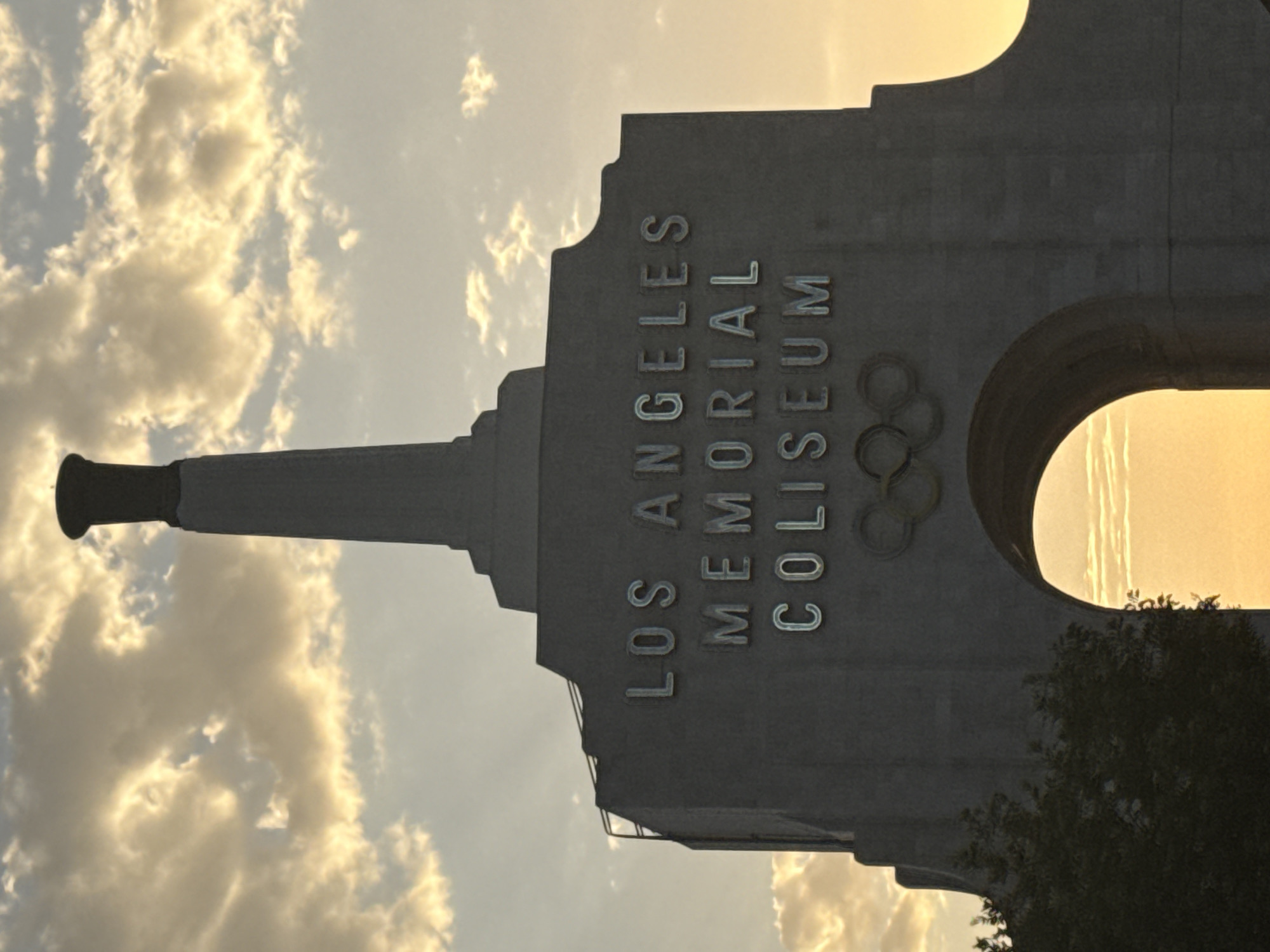 The image shows the Los Angeles Memorial Coliseum at dusk. The iconic structure is silhouetted against a sky filled with clouds illuminated by the setting sun, casting a warm, golden glow. The words "LOS ANGELES MEMORIAL COLISEUM" are prominently displayed in large, metallic letters across the facade of the building. Below the lettering, the Olympic rings symbol is visible. To the left of the structure, a silhouetted tree is partially visible. The archway of the Coliseum is in the foreground, revealing the bright, hazy sky beyond.