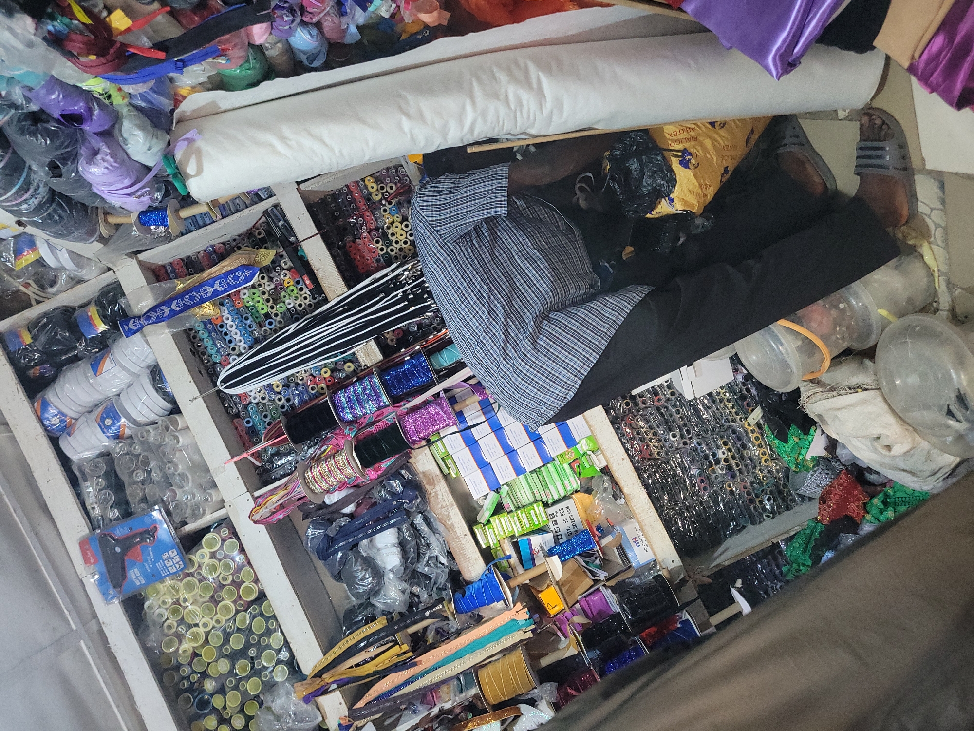 The image captures a cluttered scene within a fabric or sewing supply store. The primary focus is on the shelves packed with various sewing notions. Rolls of fabric, spools of thread, ribbons, zippers, buttons, and other crafting materials are visible. A person is seated among the shelves, wearing a checkered shirt and dark pants. Plastic storage containers, and possibly discarded packaging, are scattered nearby. The location is a dense, disorganized retail environment, typical of small businesses in Unknown, Unknown.