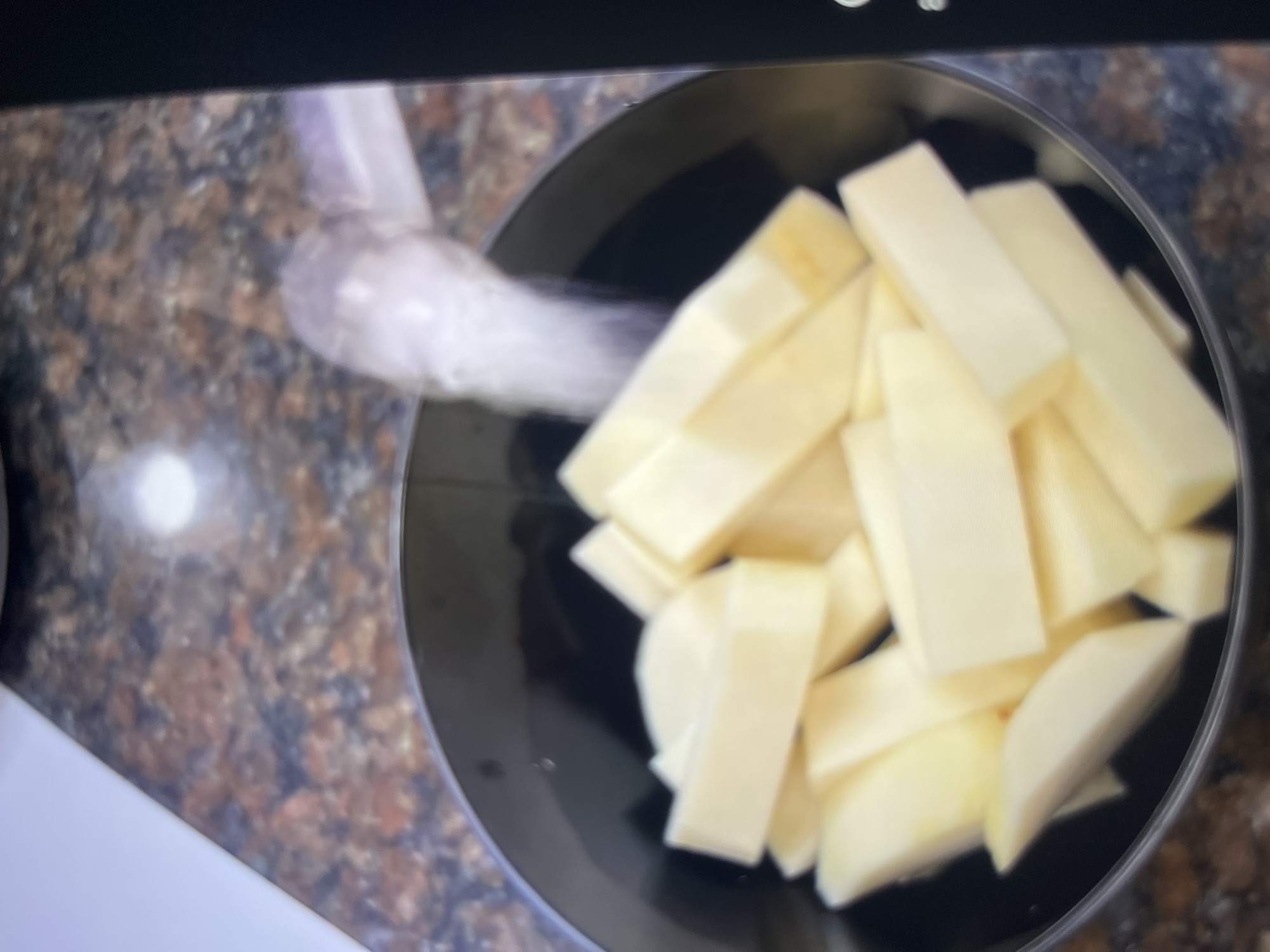 A black bowl filled with cut potato wedges sits on a speckled granite countertop. Water is being poured from a clear container into the bowl, creating a misty spray.