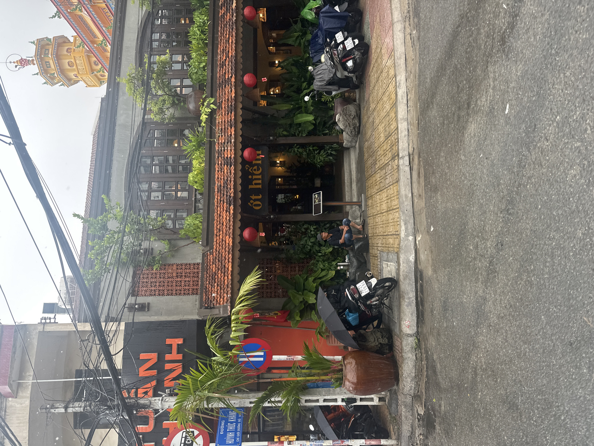 The image depicts a street scene in Nha Trang Ward, Vietnam. "OT HIEN" restaurant is visible, adorned with red lanterns. Motorbikes are parked along the sidewalk. A man is resting on the curb near the entrance. A sign indicating "HUỲNH THÚC KHÁNG" street is visible. Overhead, a complex web of power lines spans the scene. The image suggests a rainy or overcast day. A temple structure is partially visible in the upper-left corner. Plants in pots and foliage decorate the restaurant's exterior.