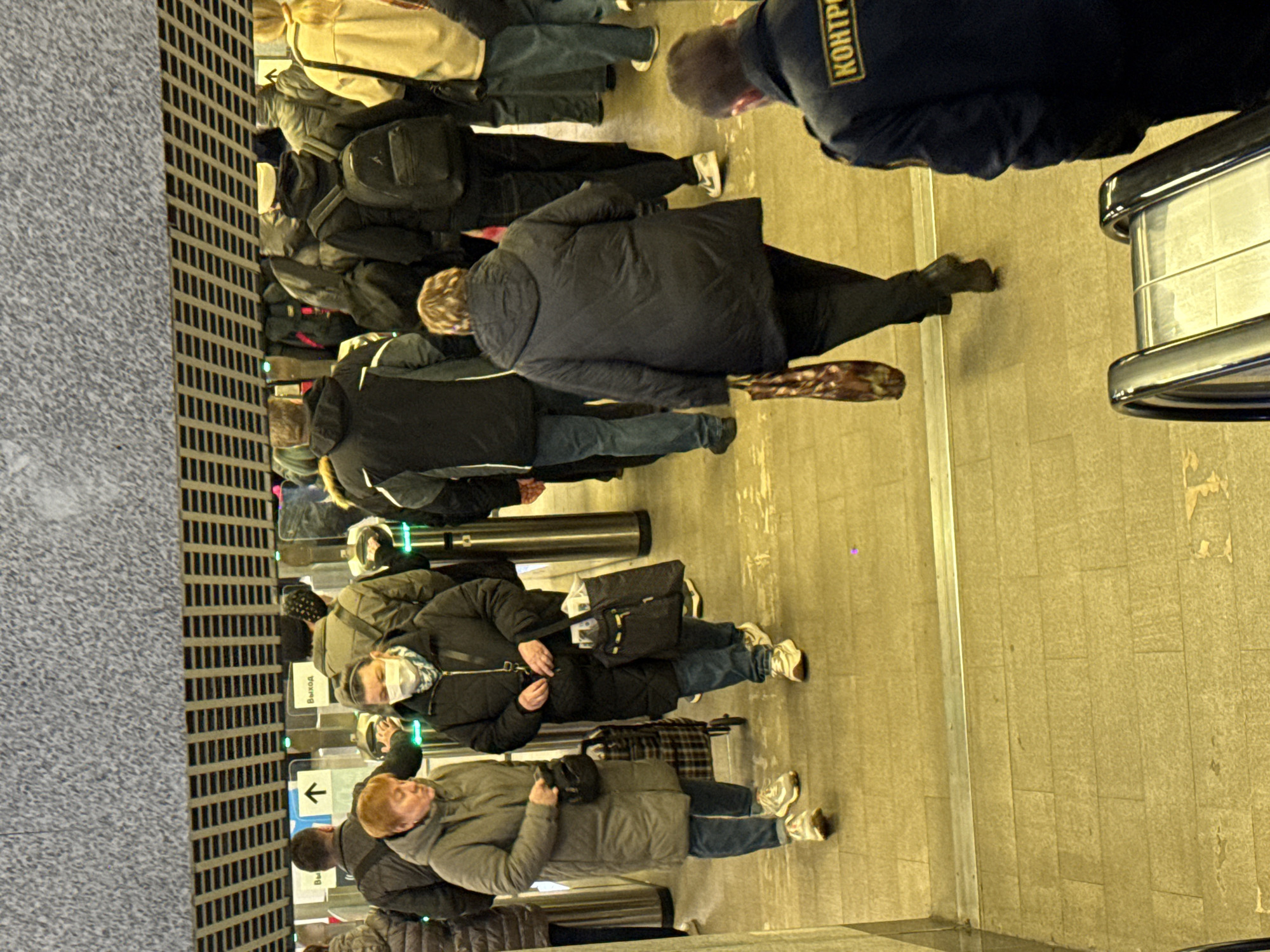 This image captures a busy indoor scene at a public transport hub, likely a subway or train station in Khimki, Russia. Numerous individuals, predominantly adults, are shown navigating a row of electronic turnstiles. They are dressed in warm winter clothing, including heavy jackets and coats, suggesting cold weather outside.
People are seen queuing and actively passing through the turnstiles, many carrying bags, backpacks, and one person with a wheeled piece of luggage, indicative of daily commutes or travel. One individual near the turnstiles is wearing a face mask. In the upper right quadrant, a person in a dark blue uniform is visible from behind, bearing the partial Russian word "КОНТРОЛ" (likely "КОНТРОЛЬ," meaning "CONTROL" or "INSPECTION") on their back, indicating a security or inspection role.
Visible text includes "ВЫХОД" (meaning "EXIT") on a sign above one of the turnstiles, and an arrow pointing left, guiding the direction of flow. The scene is illuminated by artificial yellow lighting, creating an indoor ambiance. The floor is tiled with a light-colored material, and a textured, dark gray grating runs along the left edge of the frame. The overall atmosphere is one of focused movement and routine activity typical of a public transit environment.