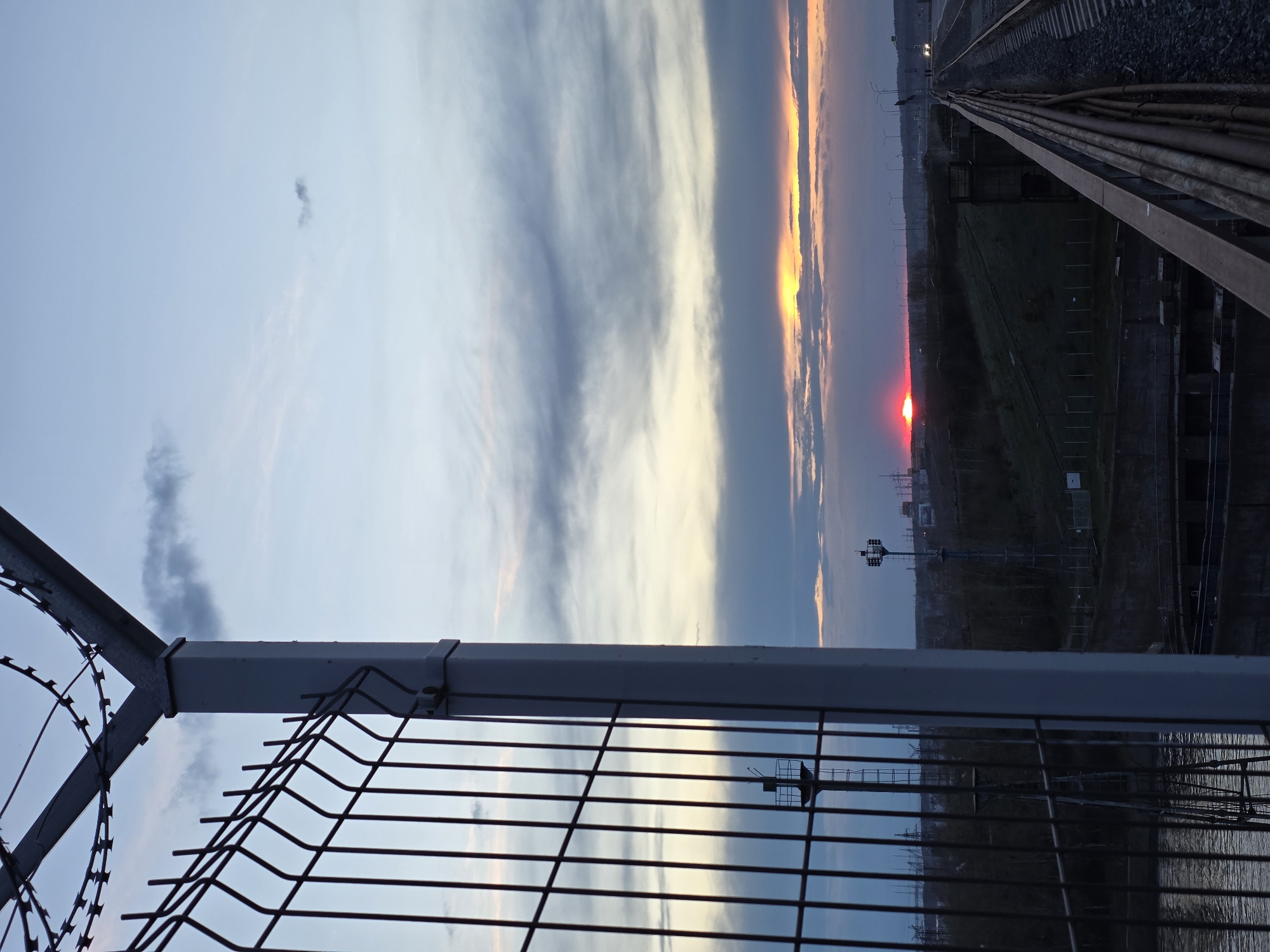 The image depicts a sunset viewed through a wire fence topped with barbed wire. A railway line stretches into the distance to the right of the frame. The red setting sun is partially obscured by clouds. Below the horizon, industrial structures, including utility poles and buildings, are visible. The fence reflects these structures. The photo was taken near Chaikovsky, Russia.