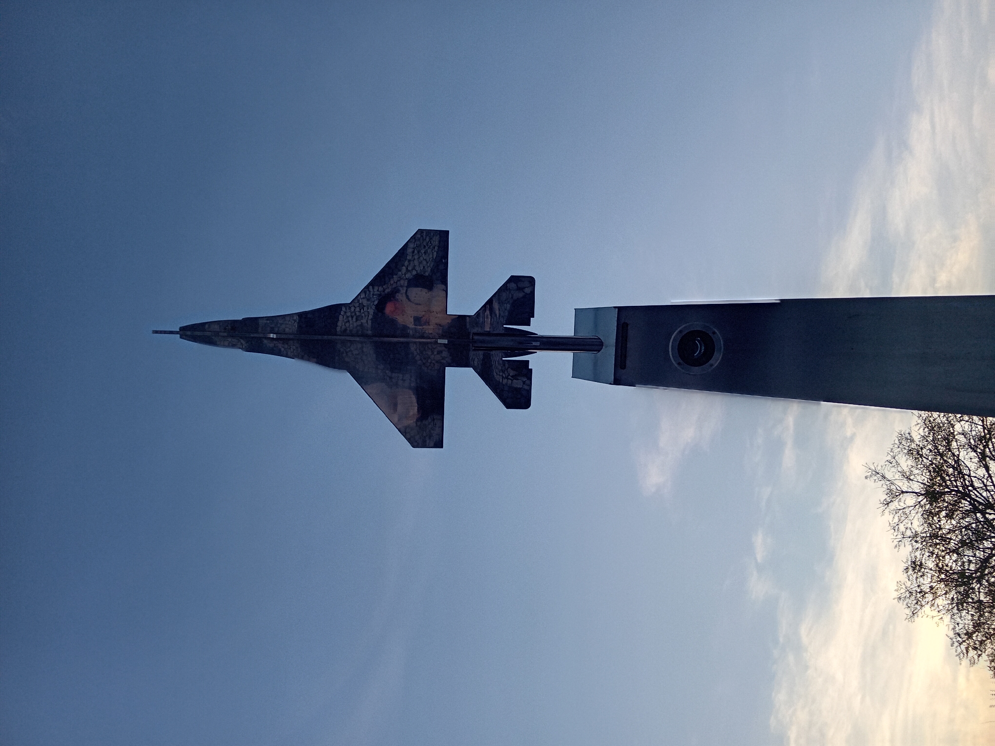 An upward-angle shot shows a McDonnell Douglas F-4 Phantom II fighter jet mounted atop a silver, rectangular, tapered pedestal. The plane faces left and the pedestal points toward the bottom-right corner. The plane is adorned with a speckled camouflage pattern and possibly a faded image on the upper surface of the wing. In the background is a clear, blue sky with sparse white clouds in the upper-right corner. A tree with bare branches is partially visible in the lower-right corner. The location is Unknown, Unknown.