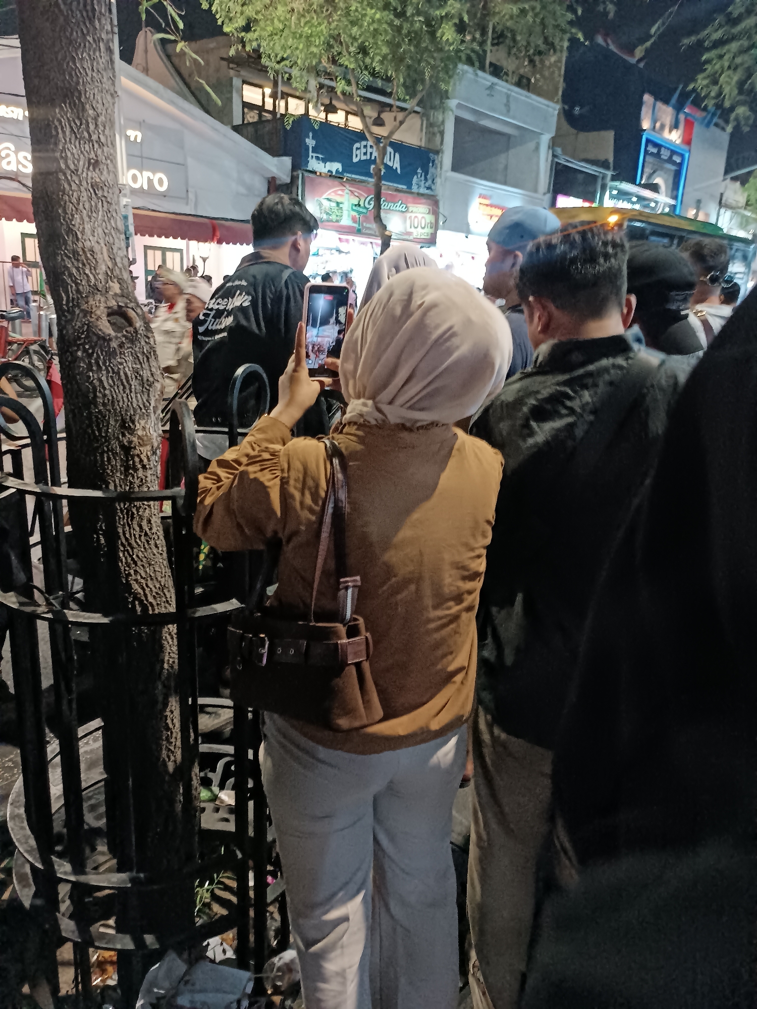 The image captures a nighttime street scene in Yogyakarta, Indonesia, likely near Malioboro Street, a well-known tourist area. A woman wearing a beige headscarf and a brown jacket is the central figure, holding up a smartphone to take a photograph. She is wearing light-colored trousers and a brown shoulder bag with a buckle detail.
In front of her, a man in a black t-shirt with white text that reads "Aceh" and "Fullo" is visible from the back, with his hair cut short. To the right of the woman, a man in a dark jacket and a blue cap is partially visible. Further back, other individuals are seen, some with dark clothing, and a blurry vehicle, possibly a bus or van, is present.
The background reveals the facade of shops with illuminated signs, including one that reads "GEF" and another with "100rb" and "3 pcs," suggesting commercial activity. A large tree trunk stands to the left, partially obscuring the view, and a black metal fence surrounds it. The ground is littered with some debris. The overall atmosphere suggests a busy, public space at night, possibly during an event or market.