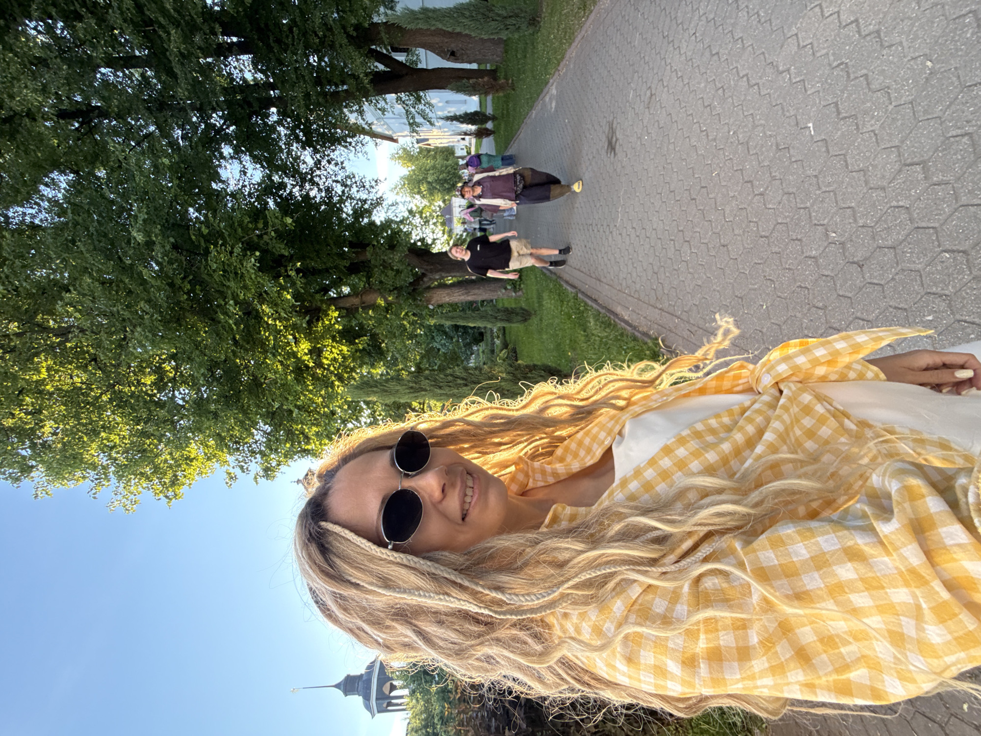 The image depicts an outdoor scene in what is identified as the city of Rusanovo, Russia, under clear blue skies with warm, golden hour sunlight. In the foreground, an adult female with long, wavy blonde hair, styled with some braided strands, faces the viewer. She wears dark oval-shaped sunglasses and a faint smile. Her attire consists of a white top or dress layered with a yellow and white checkered long-sleeved shirt, which is tied at her waist. Her fingernails are painted a light, neutral color. The sunlight creates a bright halo effect around her hair on her right side.
The foreground woman is positioned on a grey paved pathway composed of hexagonal blocks. This path extends into the midground and background, flanked on both sides by green grass and mature trees with lush green foliage. The trees cast shadows, indicating a low sun angle from the left. In the midground, two individuals walk away from the viewer along the path. The first, closer to the left, is a male wearing a black t-shirt, light-colored shorts, and dark shoes. Behind him and slightly to the right, a second individual, possibly female, wears a dark top, dark wide-legged trousers, a light-colored hat, and bright yellow slip-on sandals, carrying a dark fanny pack or belt bag. Further in the background, a small group of less distinct figures also walks along the path, some appearing to wear head coverings.
On the far left in the background, a dark-domed structure with a spire and cross, resembling a church tower, is partially visible above the trees. On the right side of the path, several multi-story white buildings with dark roofs are visible, interspersed with smaller, conical evergreen trees. The overall environment suggests a well-maintained park-like or monastic setting.