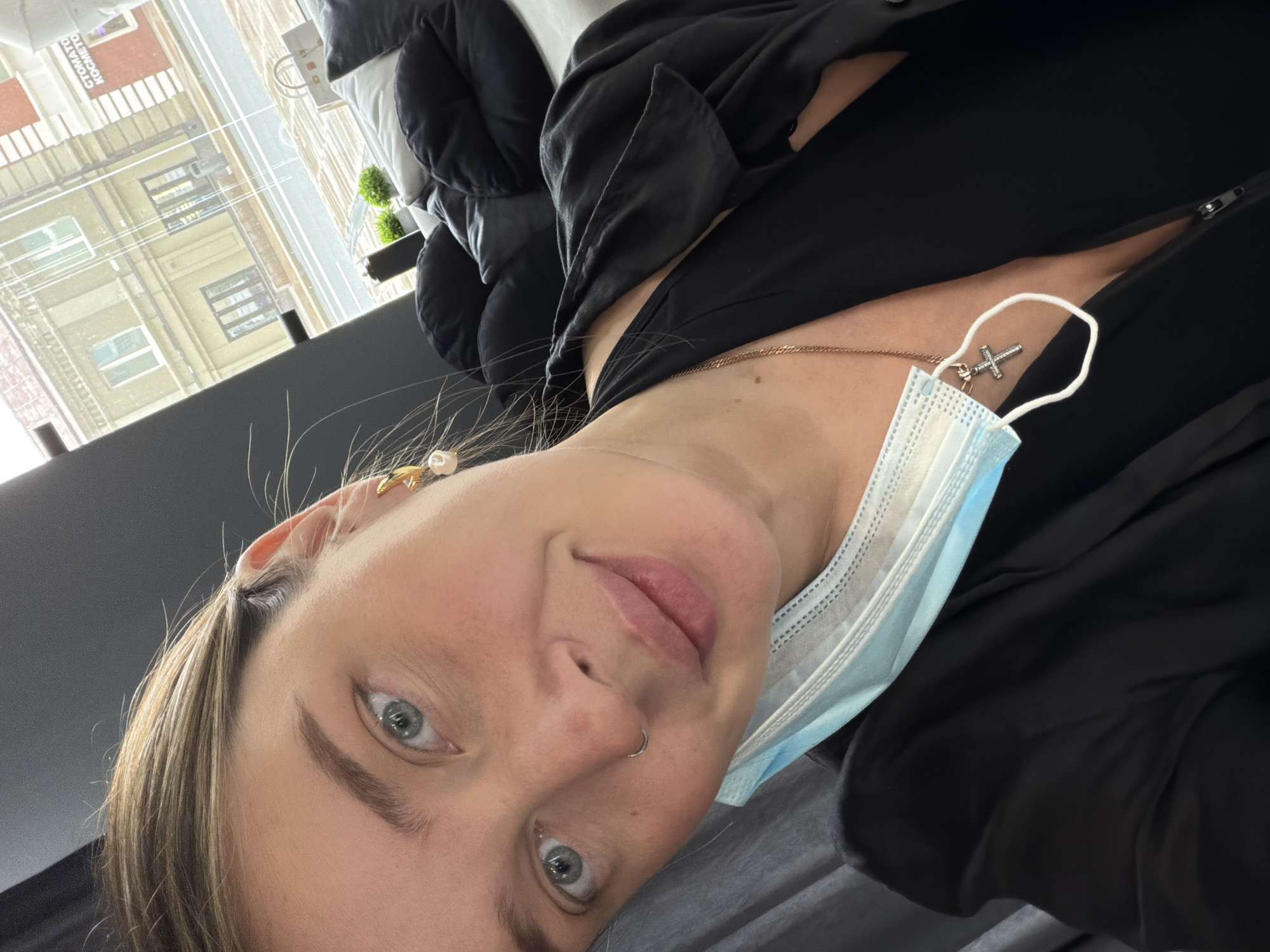 A woman is shown in a close-up, tilted, selfie. She has light skin, blue eyes, and blonde hair. She is wearing a surgical mask that is light blue and white. She is also wearing a gold chain necklace with a cross pendant. The woman is wearing a black garment. The background features a window with a view of a building with many windows. Based on the architecture and the presence of Cyrillic text visible through the window, the location is likely Moscow, Russia. The environment seems to be indoors, possibly an apartment or an office.