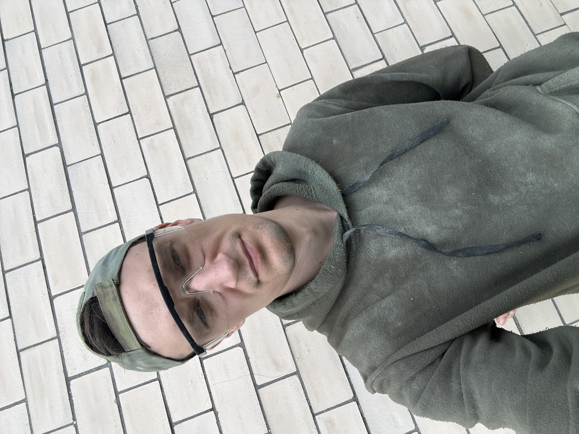 A medium shot shows a man from the chest up, wearing a green hooded sweatshirt and a green baseball cap with a camouflage pattern. He also wears clear protective glasses. The man is standing in front of a light-colored brick wall, which takes up the entire background. The wall is constructed of rectangular bricks laid in a stretcher bond pattern, with dark gray mortar. The image appears to be taken outdoors, possibly during the day, given the lighting. The man is looking directly at the camera with a neutral expression. The overall scene suggests a candid or informal portrait. The location is identified as Makiv, Ukraine.