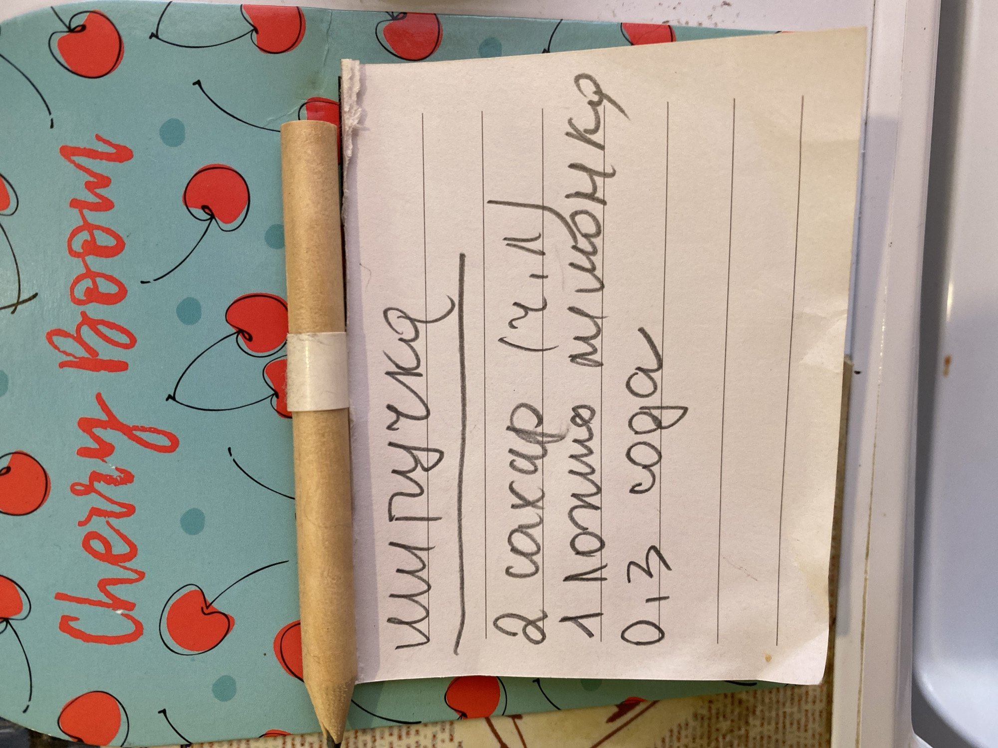 A close-up shot shows a handwritten recipe on a piece of lined paper. The paper is placed on top of a light blue notebook with a cherry pattern and the words "Cherry Boom" written in red cursive. A pencil is resting horizontally across the top of the paper. The recipe itself is written in Russian and appears to be for a beverage or dessert. The ingredients listed are: "2 саxар" (2 sugar), "1 ложка лимонно" (1 spoon lemon), and "0,3 сода" (0.3 soda). The handwriting is legible, and the scene is lit from above. The image does not provide any information about its location in Novaya Usman', Russia, or details about people, actions, or interactions.